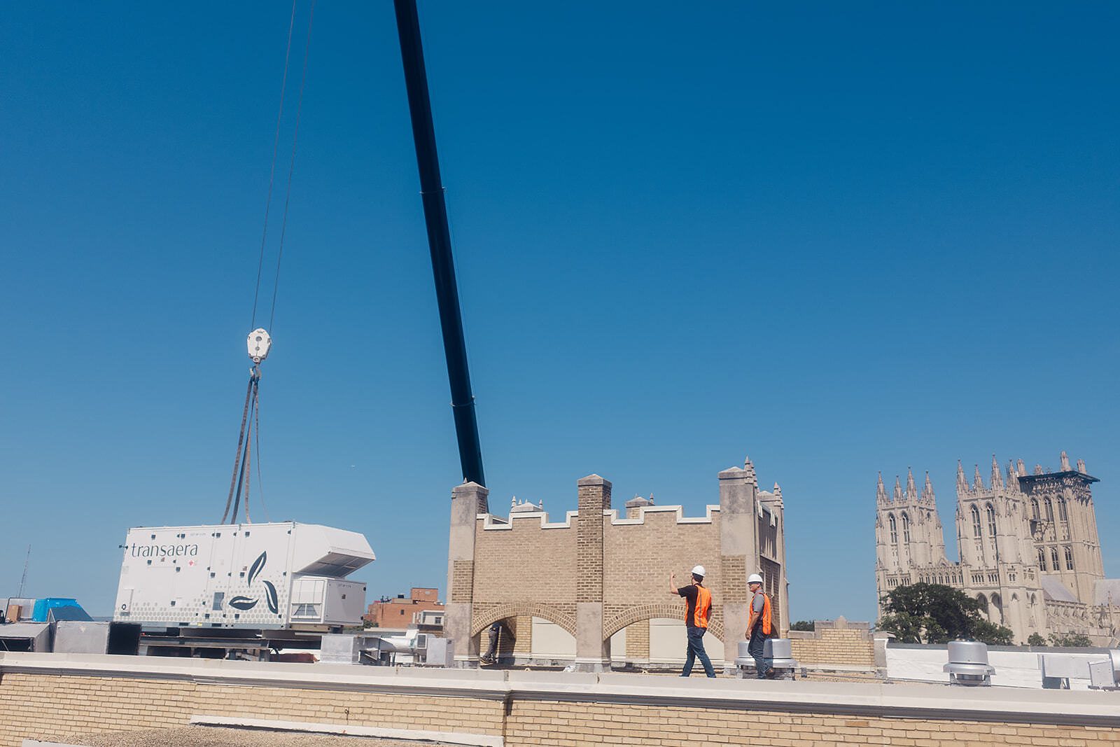 A crane hoists a white Transaera rooftop unit beside a brick building's roof parapet as two workers in hard hats and orange vests look on, with Gothic cathedral towers in the background.