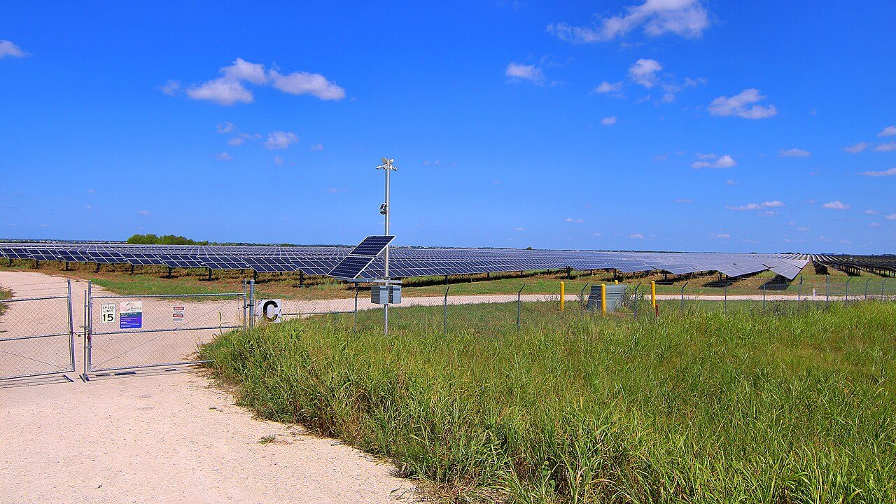 Rows of solar panels stretch behind a chain-link fence and a gated gravel road at the East Blackland Solar Project in Travis County, Texas, under a blue sky with scattered clouds.
