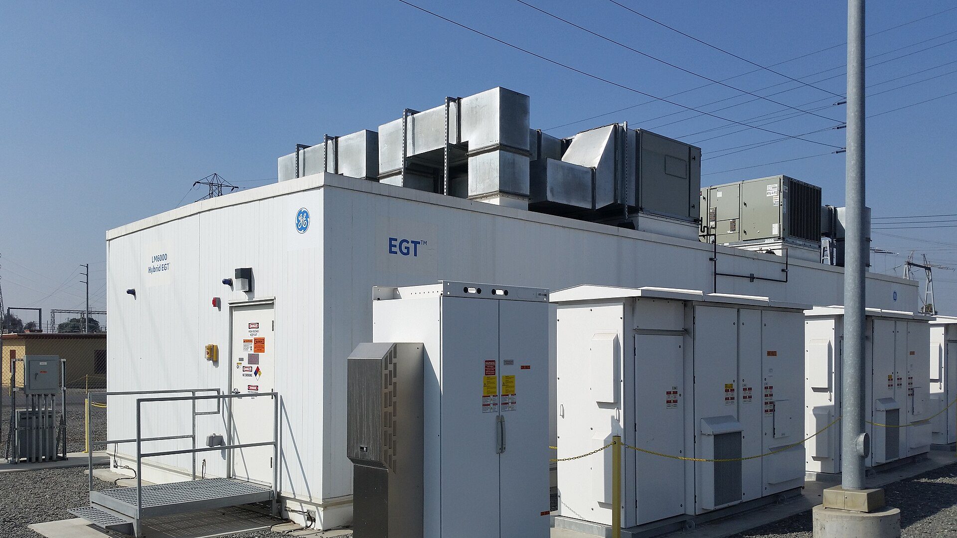 White GE enclosure marked LM6000 Hybrid EGT, with ducting on its roof and a row of white electrical cabinets in front, under transmission lines and a blue sky