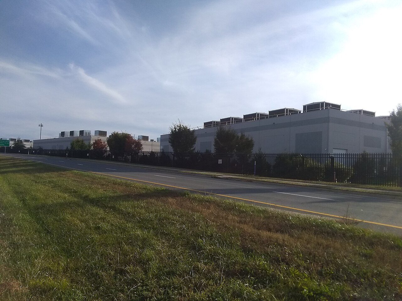 Two long, windowless gray buildings with rows of boxy rooftop equipment behind a black metal fence and trees, seen across a road and a grassy verge under a pale blue sky