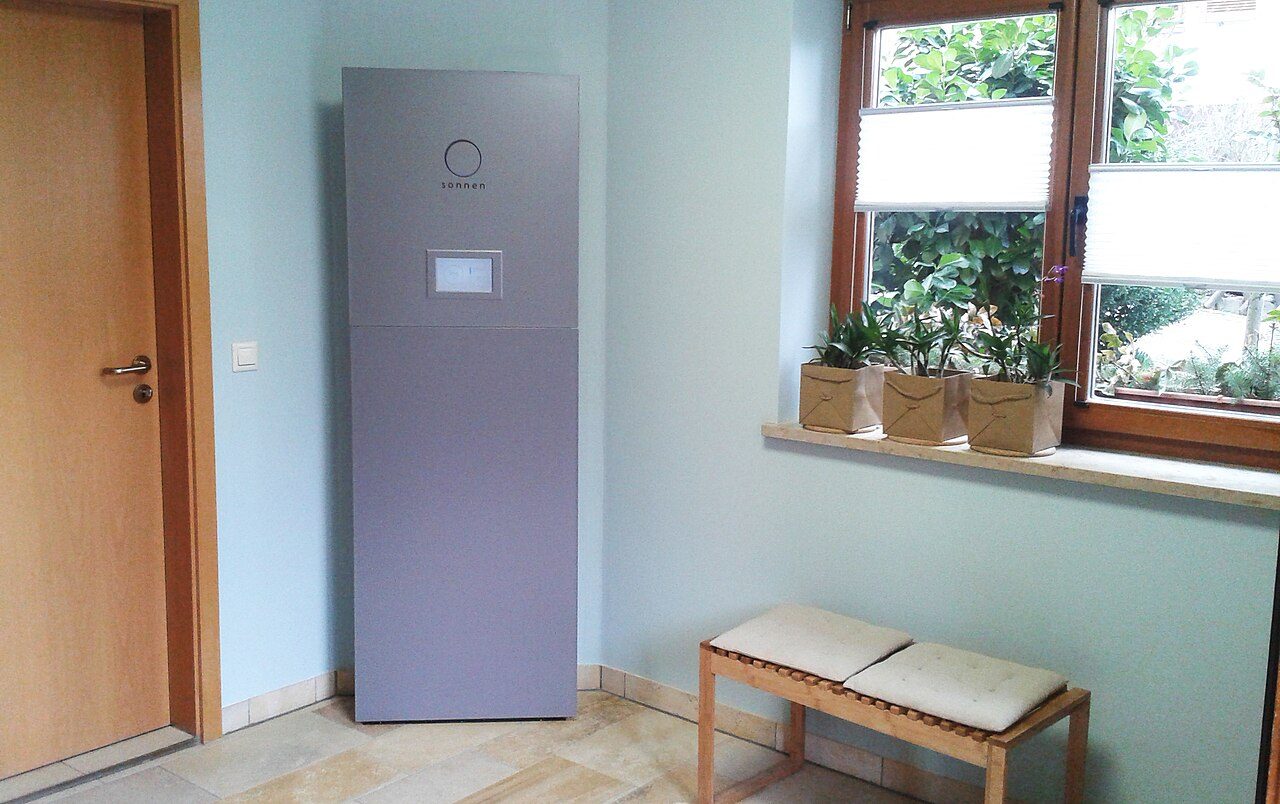 A tall grey sonnen home battery cabinet with a small display stands in a corner against a pale blue wall, beside a wooden door, a window with three potted plants and a cushioned bench.