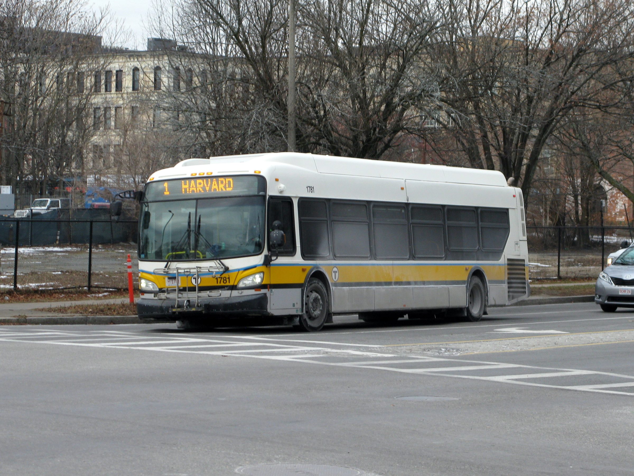 An MBTA route 1 bus signed for Harvard on Melnea Cass Boulevard at Harrison Avenue in Boston, with bare trees and a chain-link fence behind it