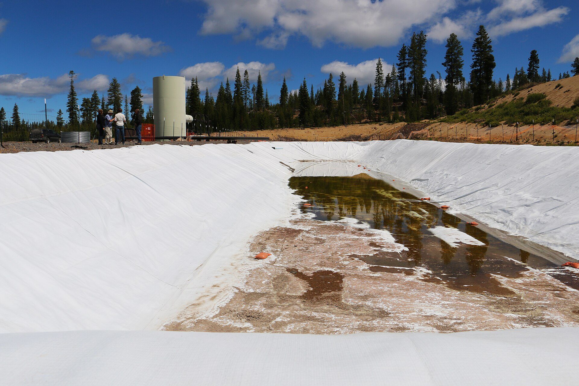 A white-lined water pond with a shallow pool at its bottom, a pale cylindrical tank and four people standing at the rim, with pine forest and a blue sky with scattered clouds behind