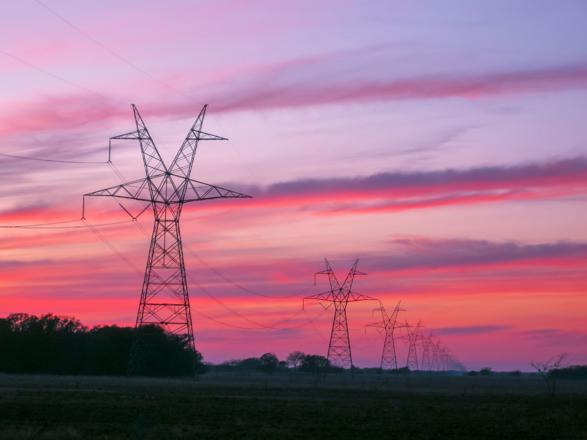 A line of steel lattice transmission towers carrying high-voltage lines across flat East Texas pasture under a pink and purple sunset sky