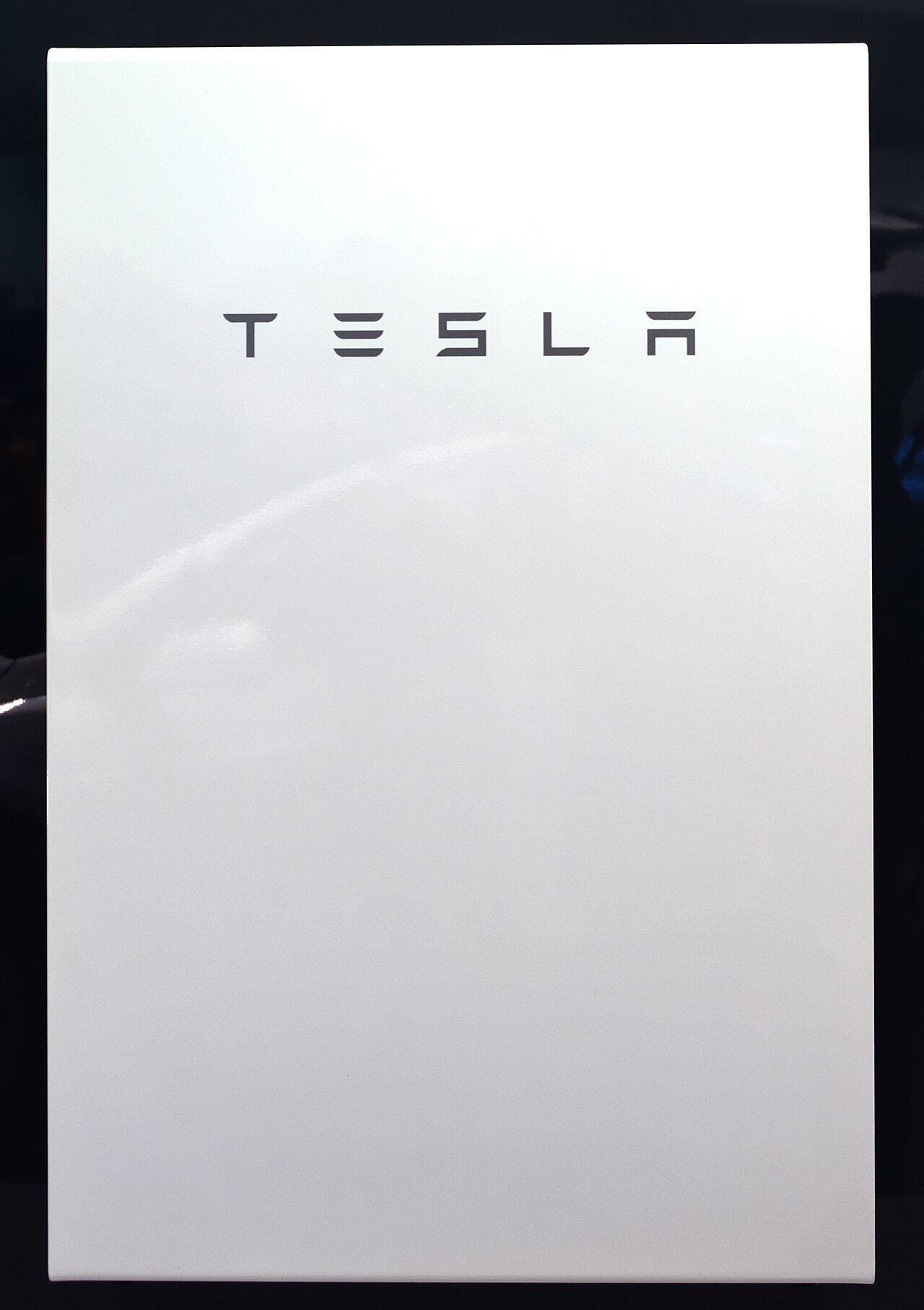A white Tesla Powerwall 2 home battery, its front panel bearing the Tesla wordmark, photographed head-on against a dark background