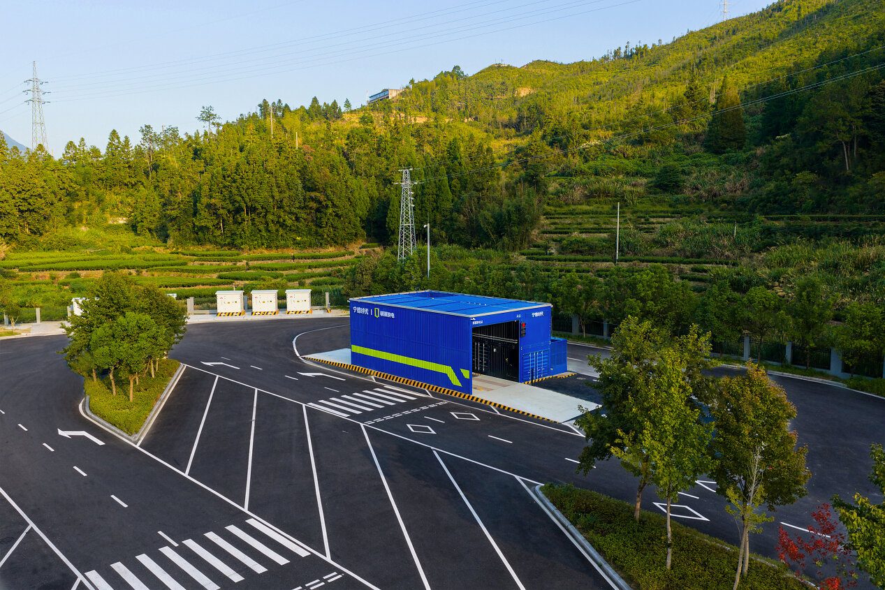 A blue Qiji battery-swap station building with an open bay beside a freshly paved, empty lot, set against forested hills crossed by power lines