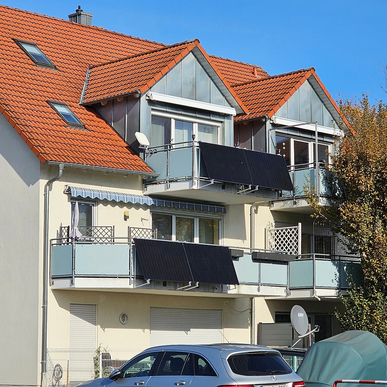Two pairs of black solar panels hung on the outside of an upper and a lower balcony railing of a cream-colored apartment building with a red tile roof under a clear blue sky, with a silver car parked in front