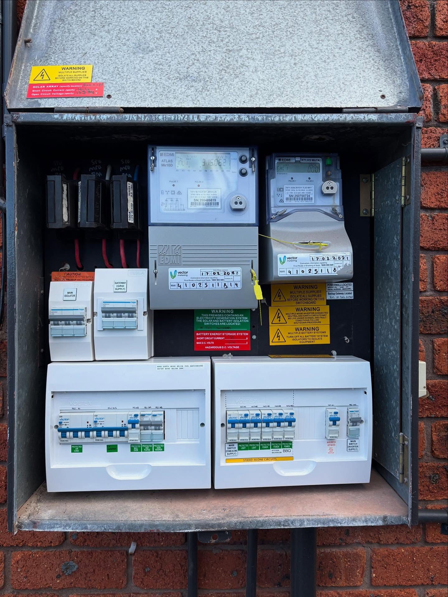 An open grey metal meter box on a red brick wall holding two electricity meters, Tesla gateway and isolator switches, white circuit breaker panels and warning labels for a solar array and a battery energy storage system