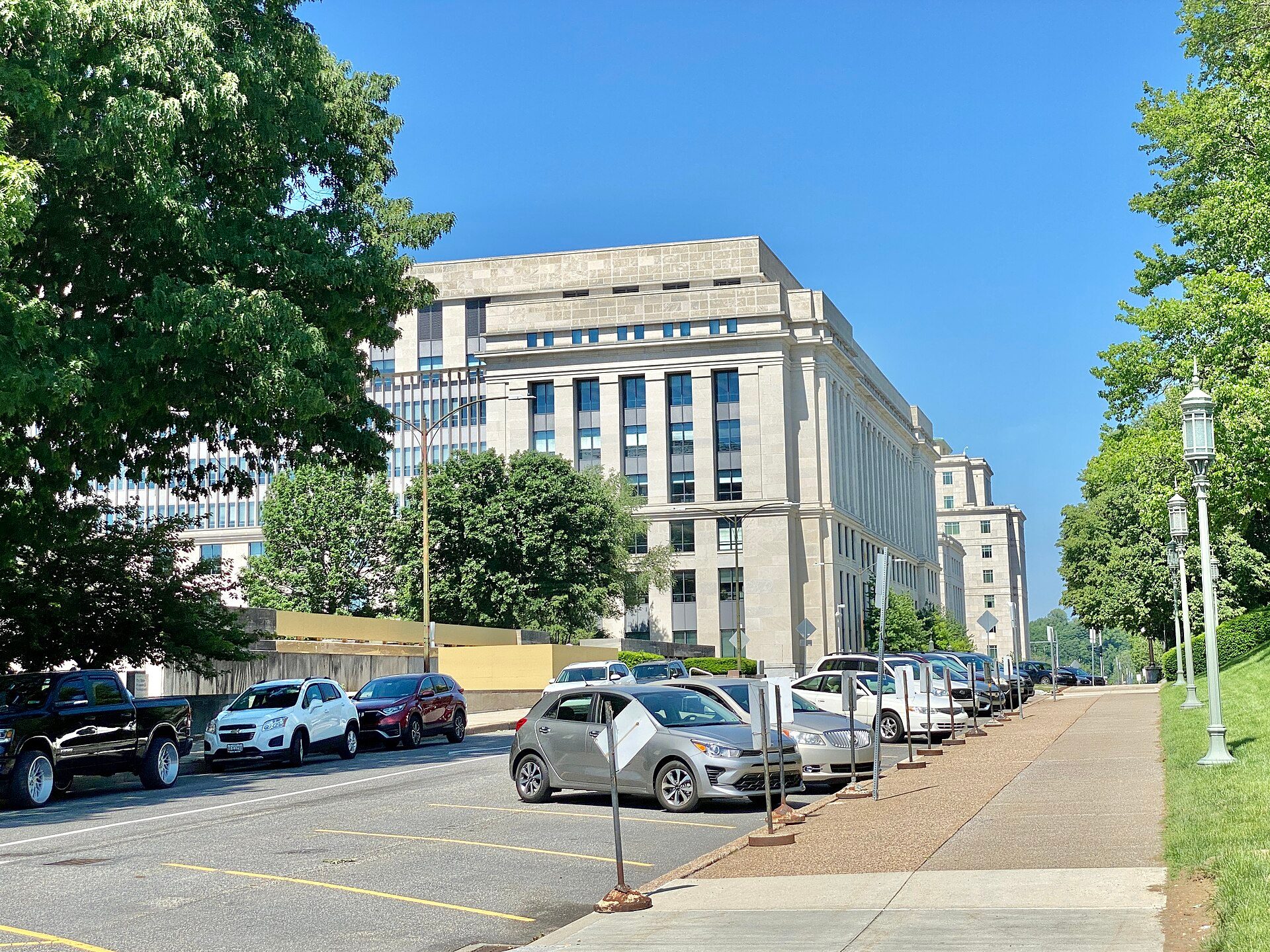 A pale stone government office building in Harrisburg, seen past trees and a street of parked cars under a clear blue sky.