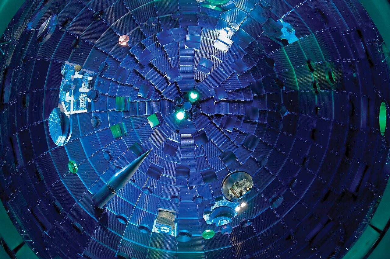 Interior of the National Ignition Facility's spherical target chamber lit blue, with the pointed target positioner extending into the chamber from the lower left and square beam ports lining the wall
