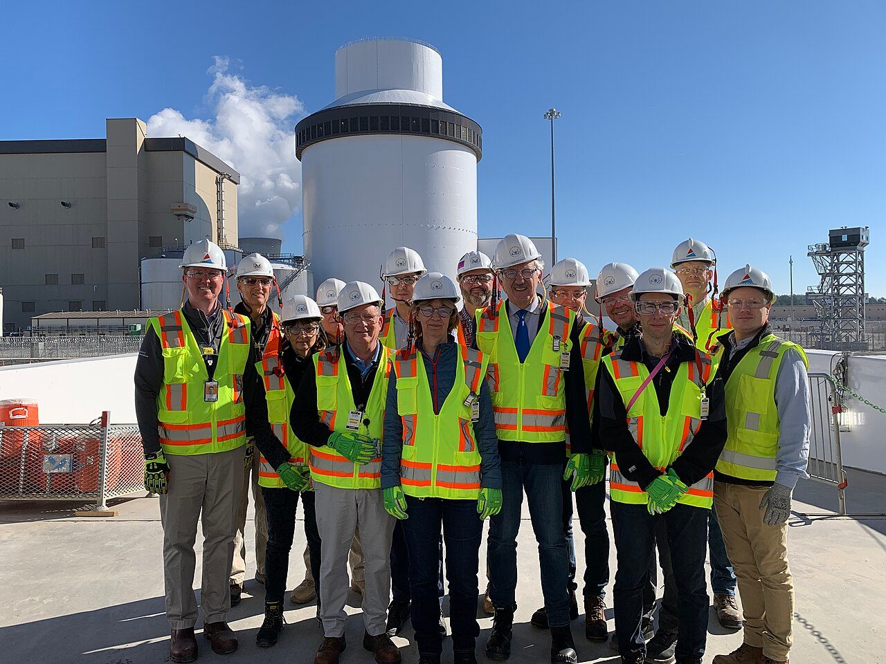 A group of about fifteen people in white hard hats and yellow high-visibility vests stands on a concrete deck in front of a tall white cylindrical reactor shield building topped by a narrower drum, with a tan turbine building and a steam plume to the left under a blue sky.