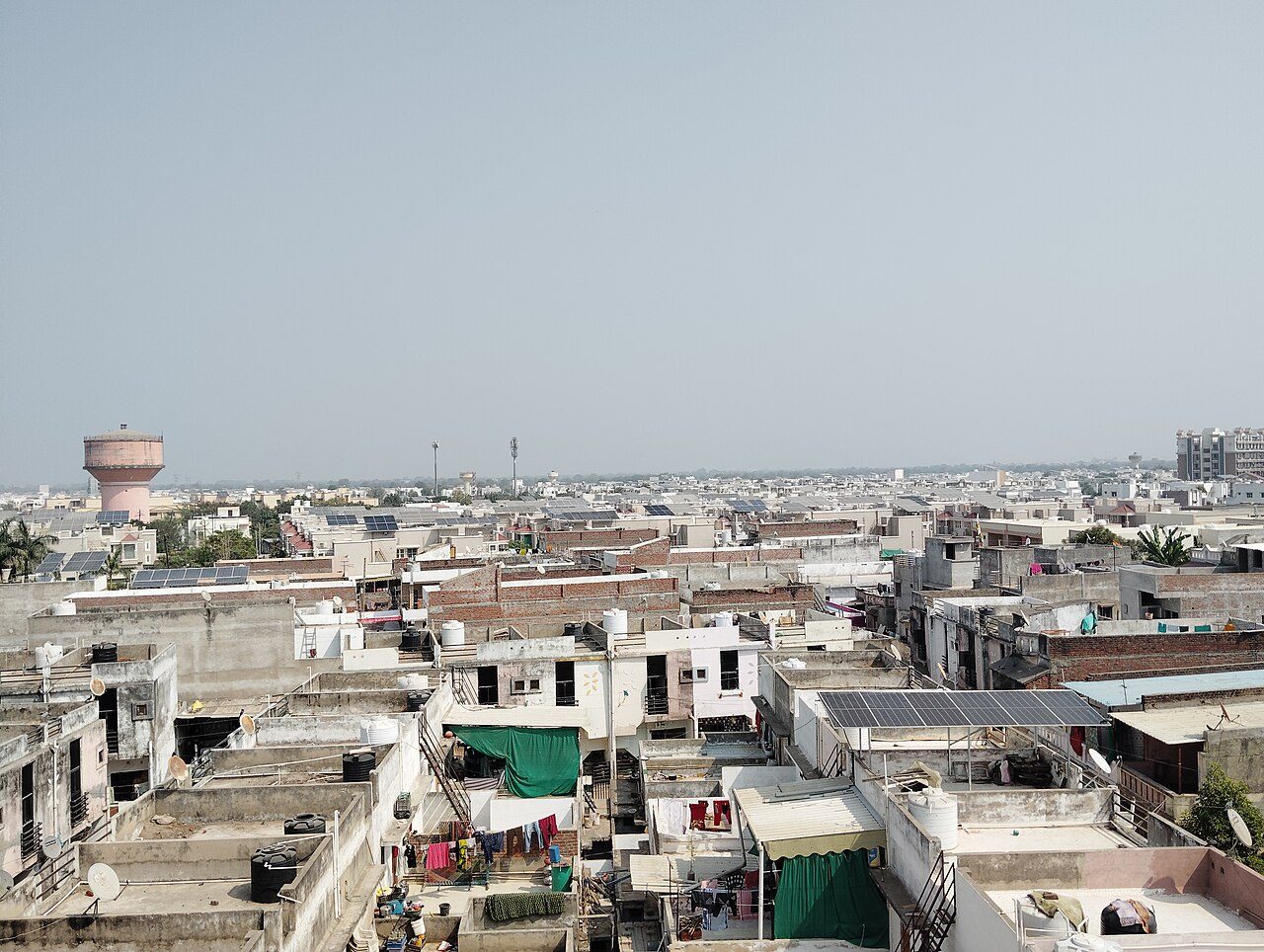 Flat concrete rooftops of homes in Mehsana, Gujarat, several carrying solar panel arrays, with water tanks and laundry on the roofs and a pink water tower on the horizon