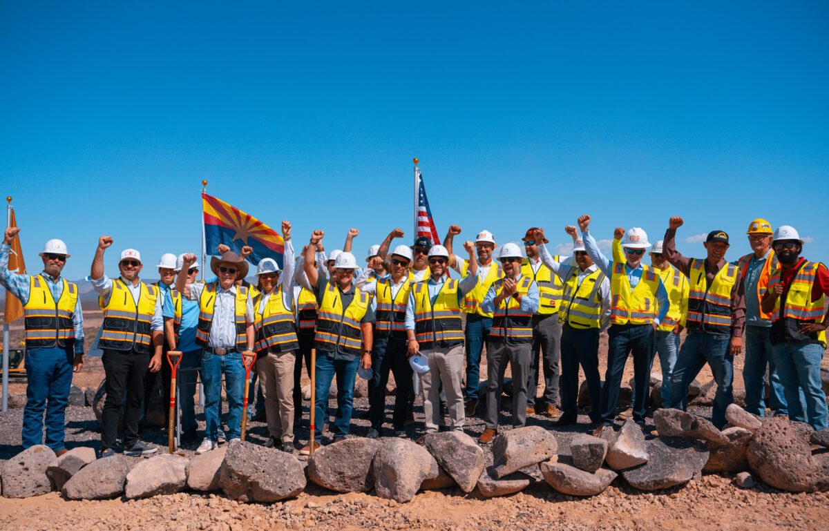 About two dozen people in hard hats and high-visibility vests raise their fists on bare desert ground behind a row of boulders, with shovels, an Arizona flag and a US flag among them