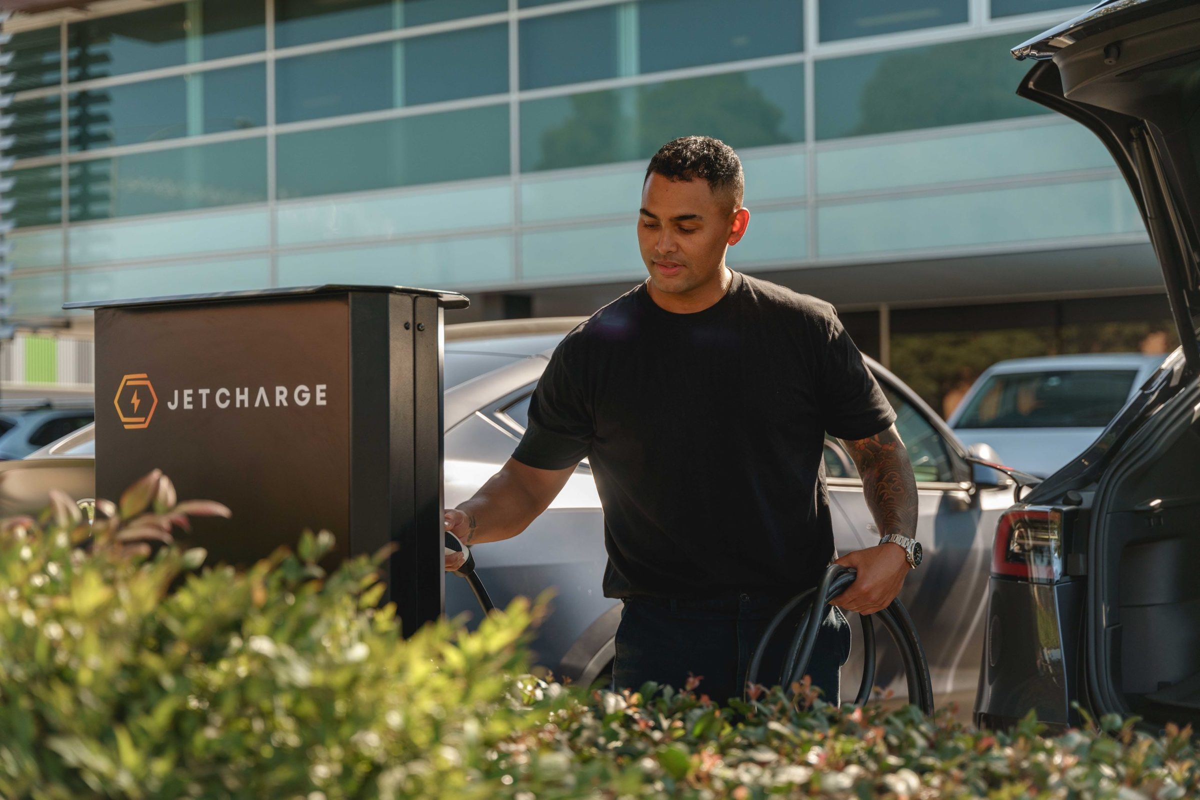 A man in a black T-shirt plugs a charging cable from a black JET Charge pedestal charger into a parked car, holding the coiled cable, with an SUV's open tailgate at right and an office building behind