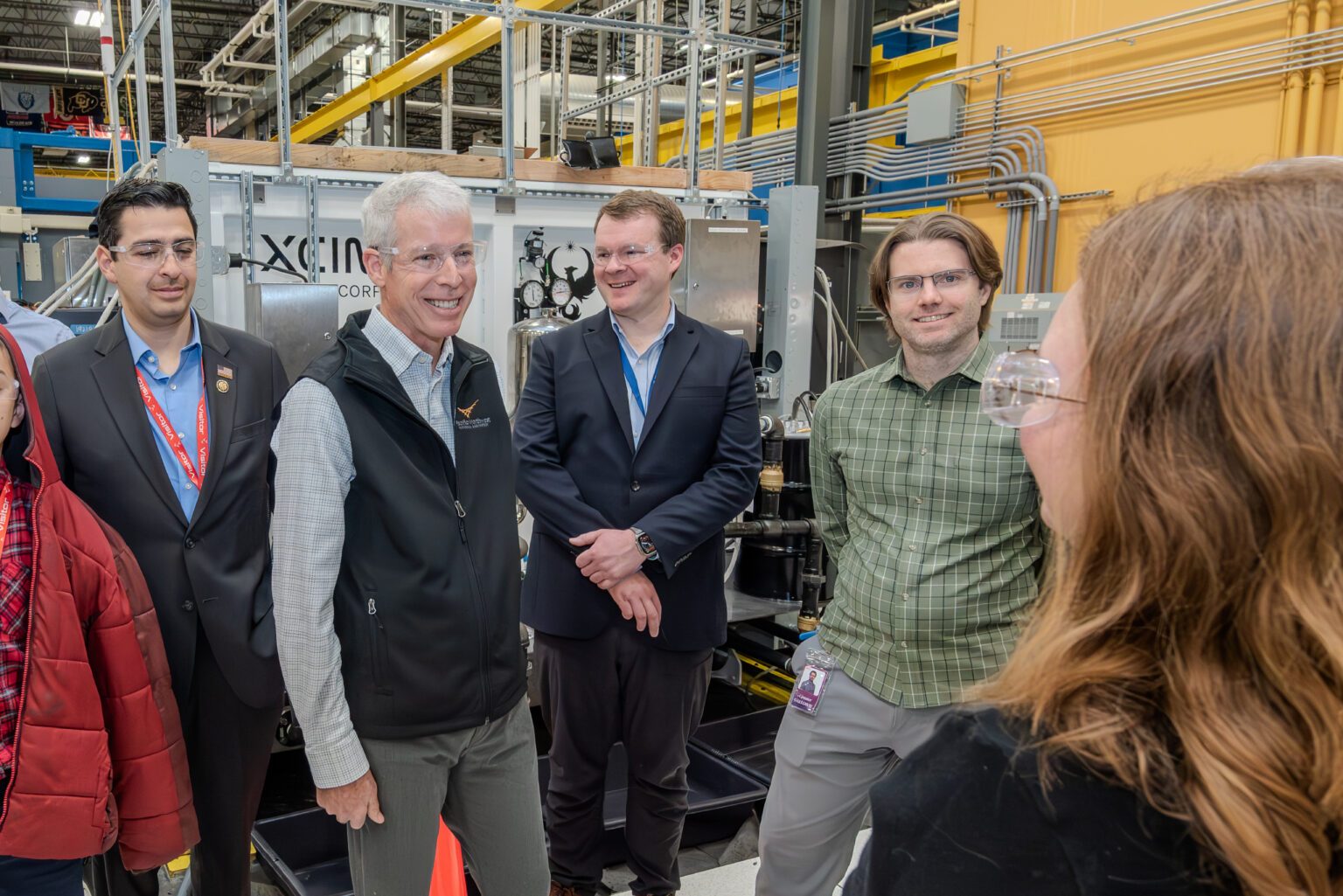 Energy Secretary Chris Wright, gray-haired in a black vest and safety glasses, smiles with a group of people in safety glasses in an industrial hall, in front of equipment with a partly visible Xcimer logo.