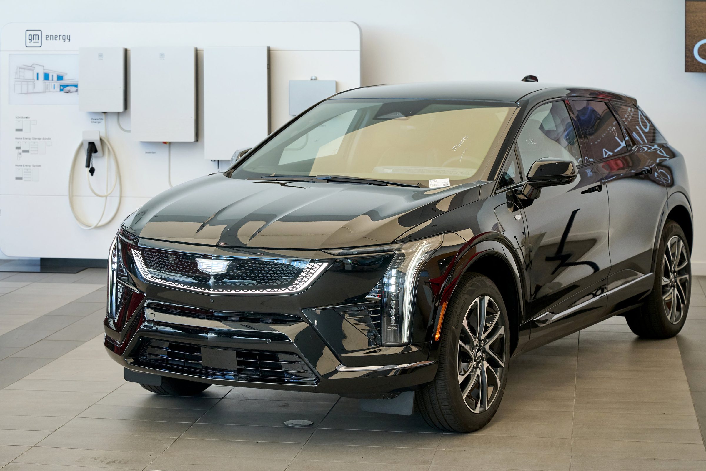 A black Cadillac-badged electric SUV parked on a tiled showroom floor in front of a white GM Energy wall display of home charger and battery units