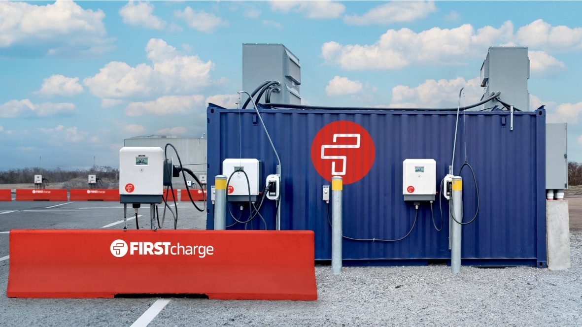 First Charge charging dispensers mounted on a blue shipping container and on posts in a paved lot, with a red First Charge barrier in front and cables running over the container roof