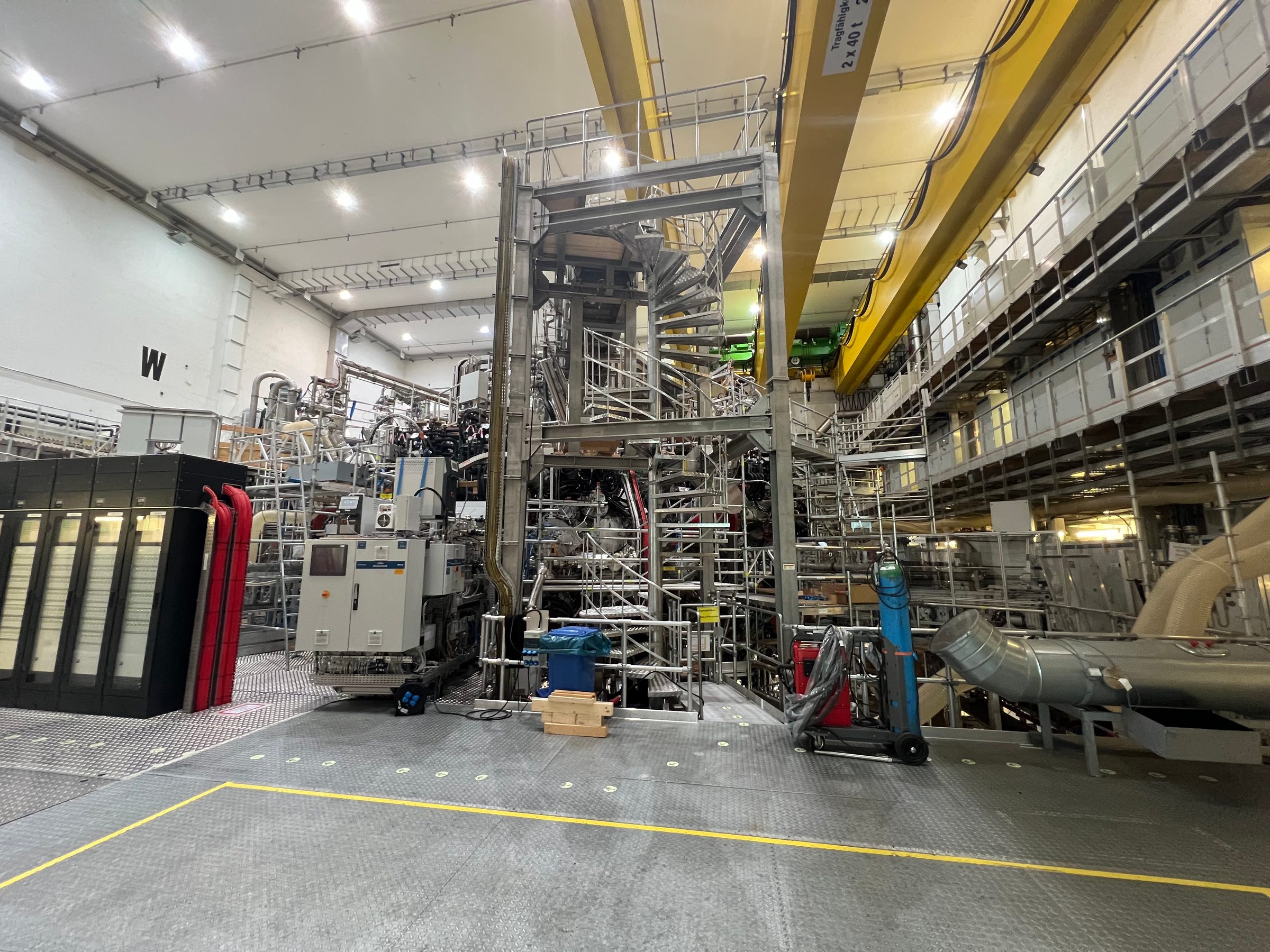 The Wendelstein 7-X stellarator hall: a tall steel frame with spiral stairs and scaffolding wrapped around the machine, pipework and equipment cabinets on the floor, and yellow overhead crane beams under a white ceiling.