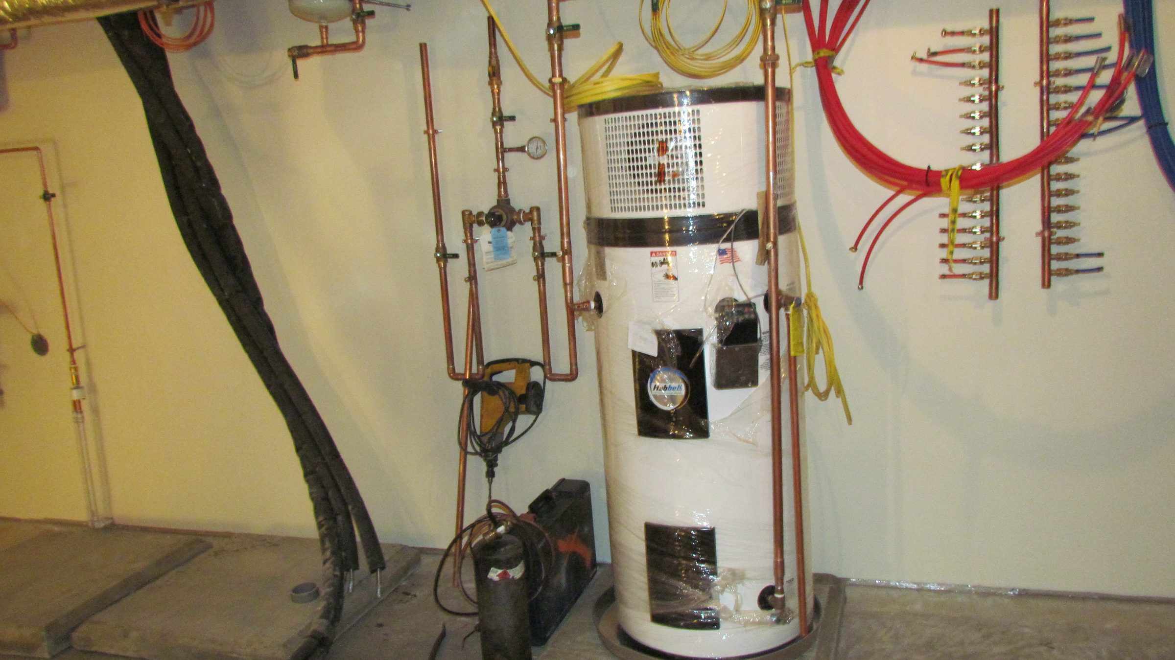 A heat pump water heater, still wrapped in protective plastic, plumbed with copper pipe in a mechanical room, with red and blue tubing manifolds on the wall and tools on the floor