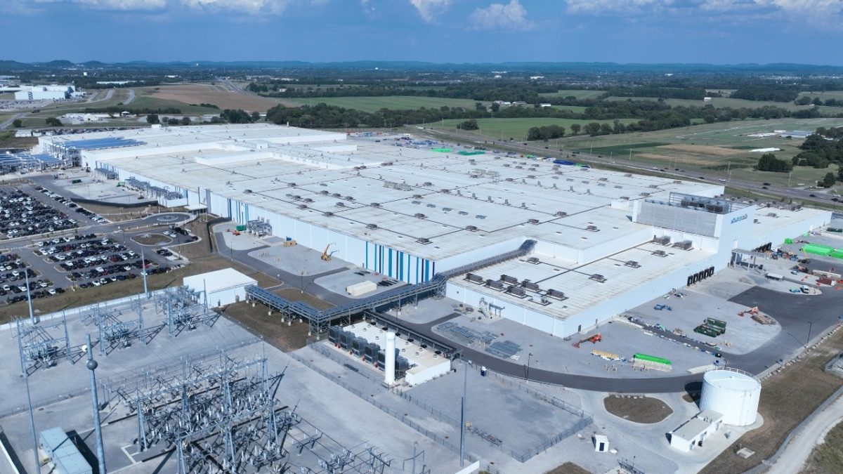 Aerial view of the Ultium Cells plant in Spring Hill, Tennessee: a large white flat-roofed factory with an electrical substation and cooling units in the foreground, a full parking lot at left, and farmland and tree lines beyond under a partly cloudy sky.