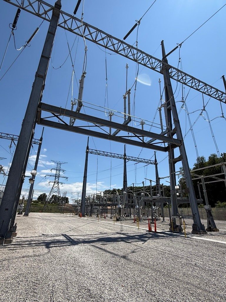 Steel switchyard structures with overhead conductors and strings of insulators rise from a gravel yard under a clear blue sky, with a lattice transmission tower and trees in the background