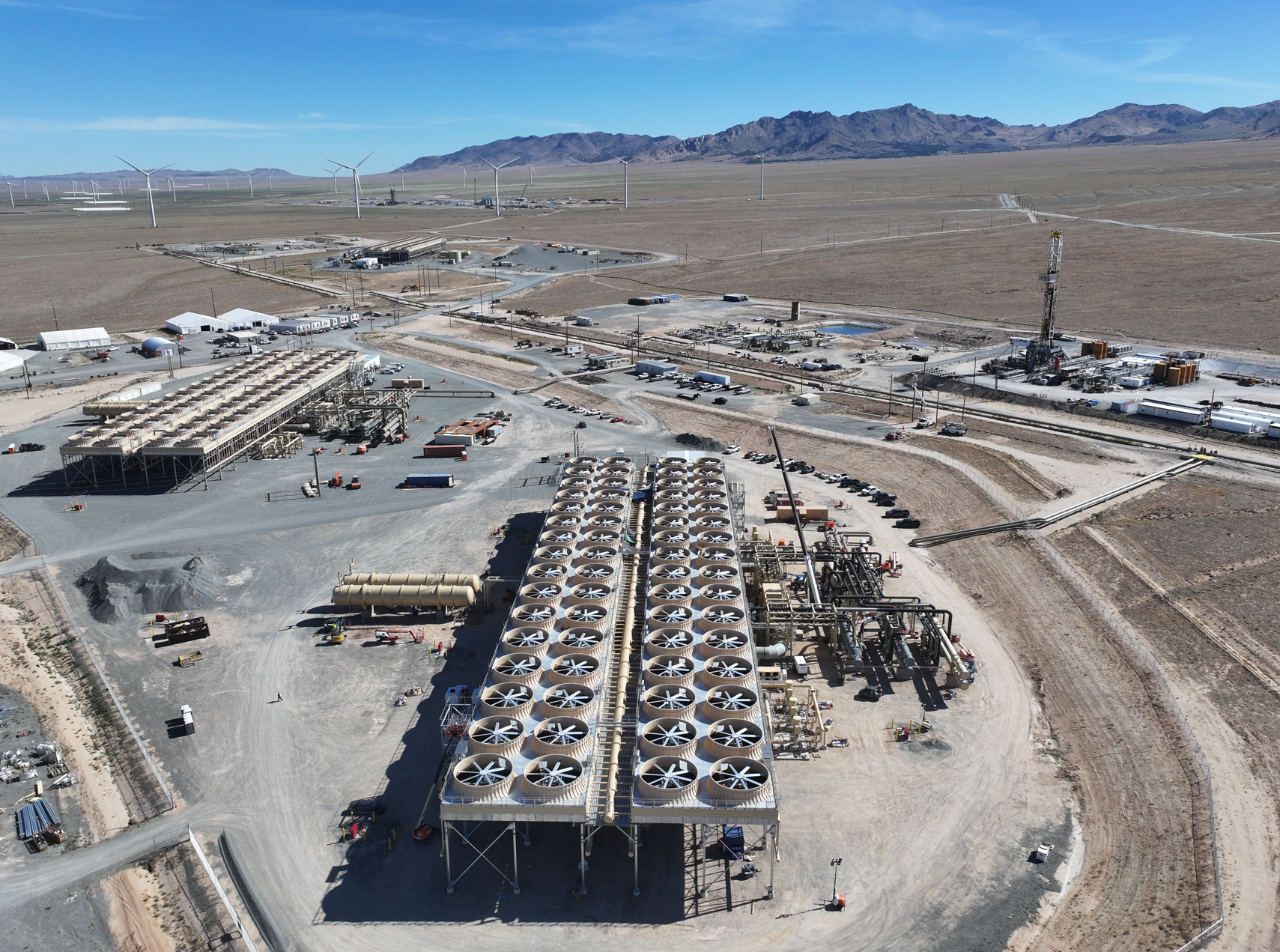 Aerial view of two banks of air-cooled condenser fans at Fervo's Cape Station geothermal plant in the Utah desert, with a drilling rig and wind turbines in the distance under a clear sky