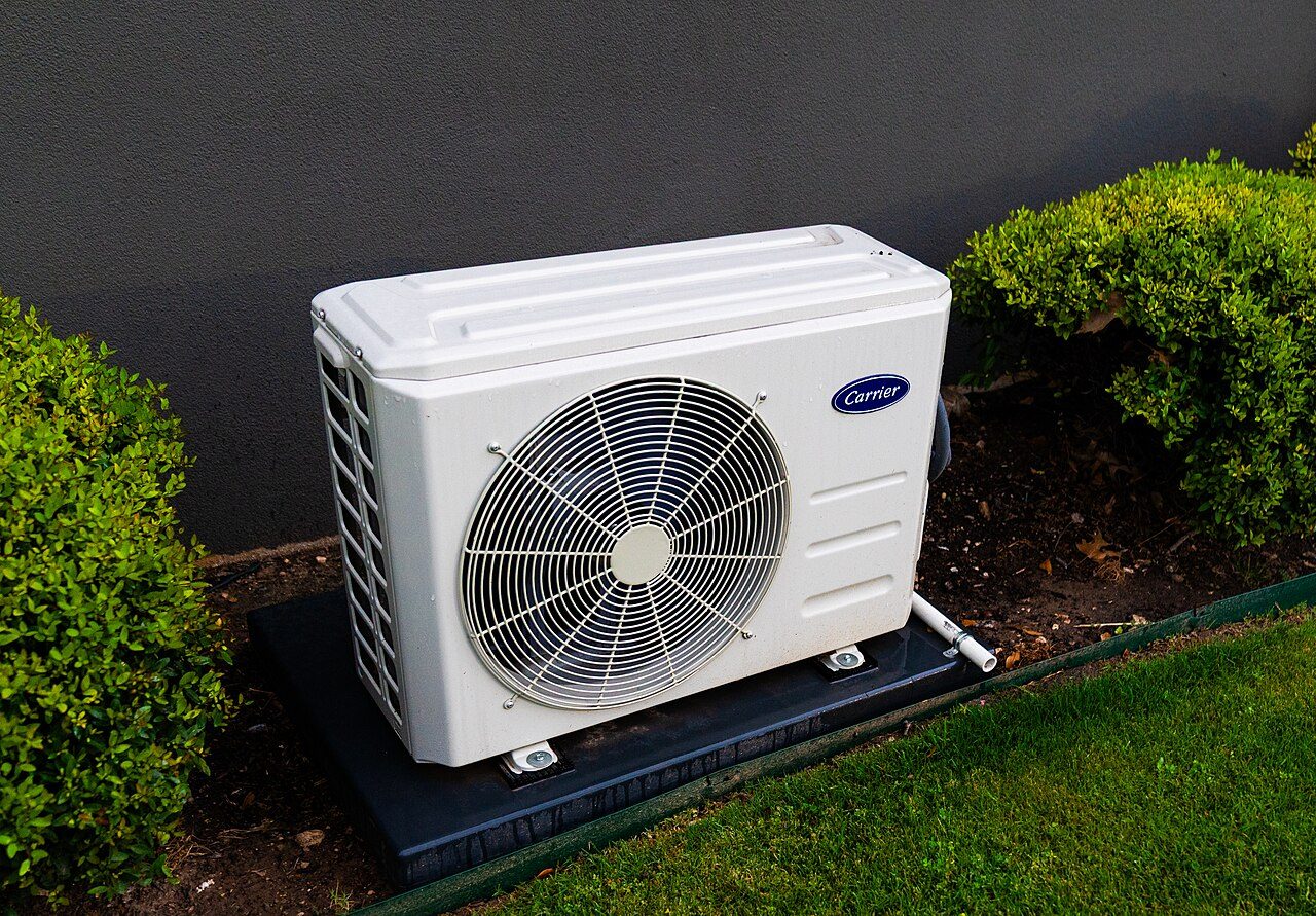 A white Carrier outdoor split-system unit with a round fan grille sits on a dark pad against a gray stucco wall between two green shrubs, with lawn in the foreground