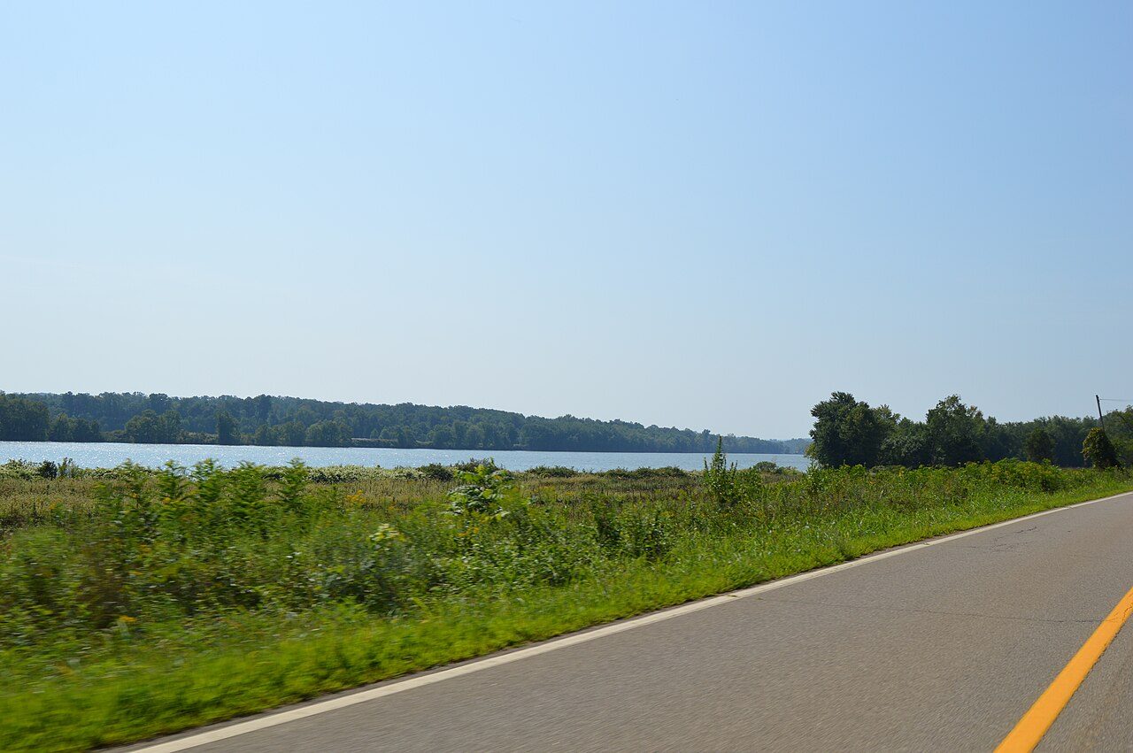 The Ohio River seen from a two-lane road in Letart Township, Ohio, with green brush in the foreground and a wooded far bank under a clear sky.