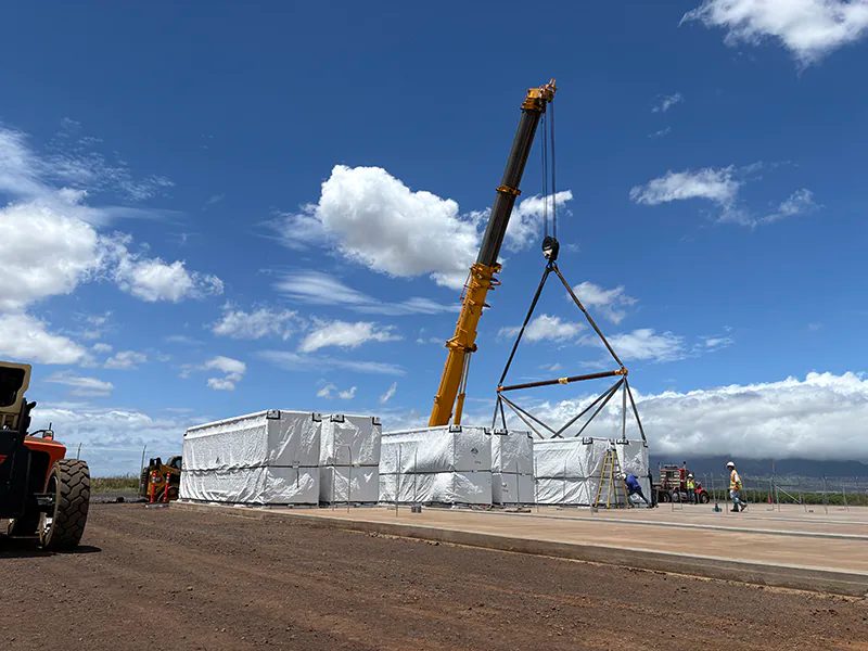 A yellow mobile crane with a rigging frame stands over a row of white-wrapped battery units on a concrete pad under a blue sky with scattered clouds; workers in hard hats stand at right and a cloud-covered ridge rises behind
