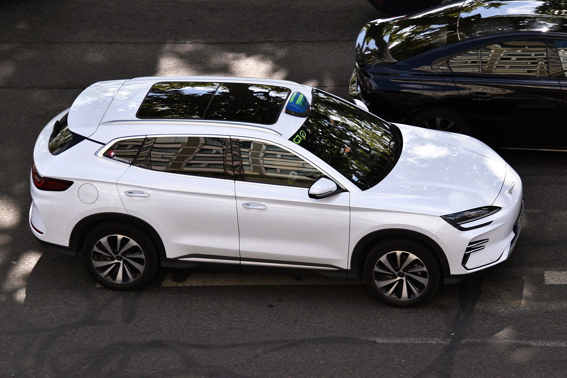 Overhead view of a white BYD Seal U DM-i plug-in hybrid SUV with a taxi roof light, stopped on a street beside a dark sedan.