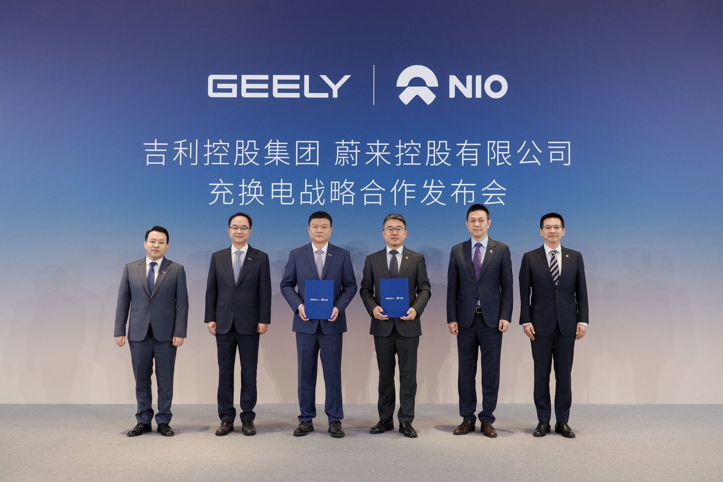 Six men in dark suits stand in a row on a stage before a blue backdrop bearing the Geely and NIO logos and Chinese text naming the two companies' charging and battery-swap strategic cooperation launch; the two at center each hold a blue folder printed with both logos.