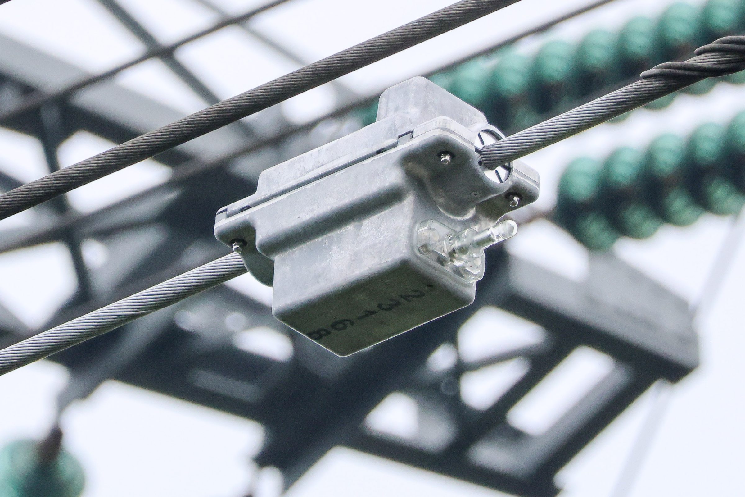 A grey metal sensor box clamped around a stranded overhead conductor, with green insulator strings and a steel lattice tower blurred behind it