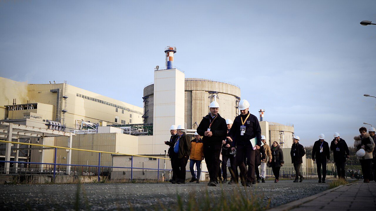 Visitors in white hard hats walk along a gravel path past a round concrete reactor building, a second smaller one and a turbine hall at Romania's Cernavodă nuclear plant.