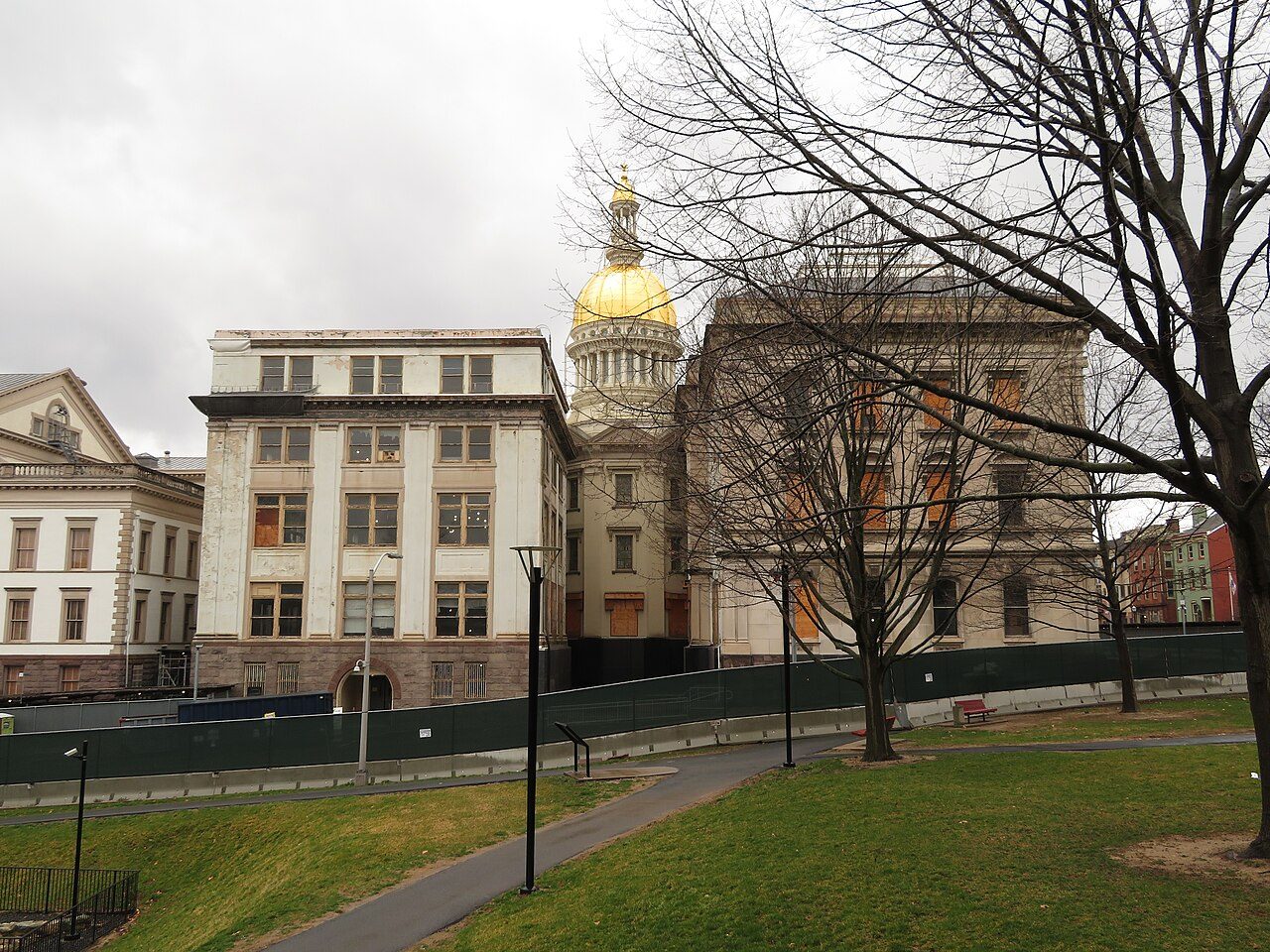 Gold dome of the New Jersey State House in Trenton rising between stone office wings, with construction fencing, bare trees and a lawn in the foreground