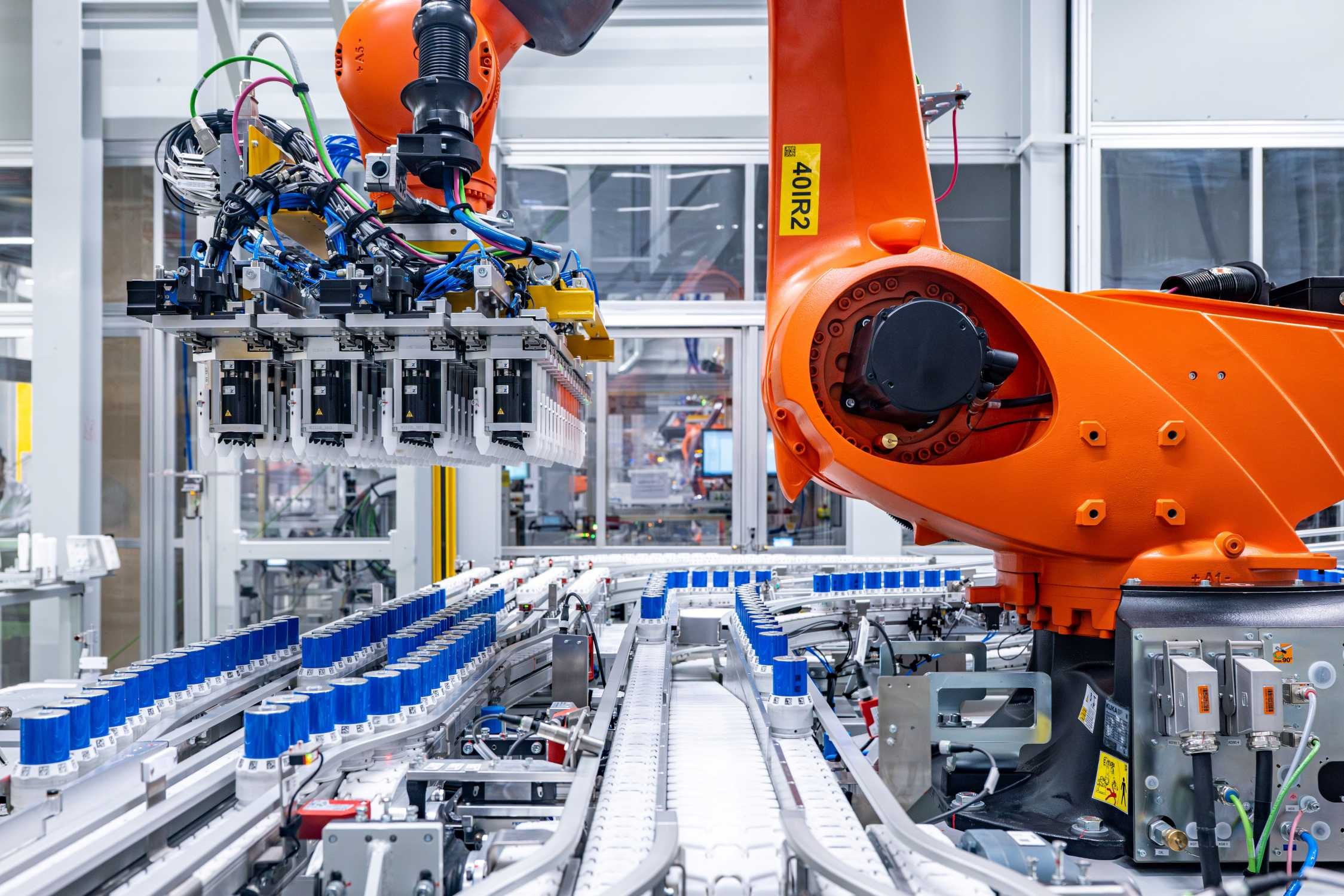 Orange industrial robot with a multi-finger gripper above conveyor lanes carrying rows of blue cylindrical battery cells