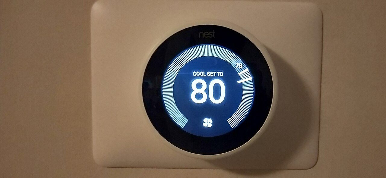 A wall-mounted Nest thermostat with a round blue display reading Cool set to 80, on a cream wall plate