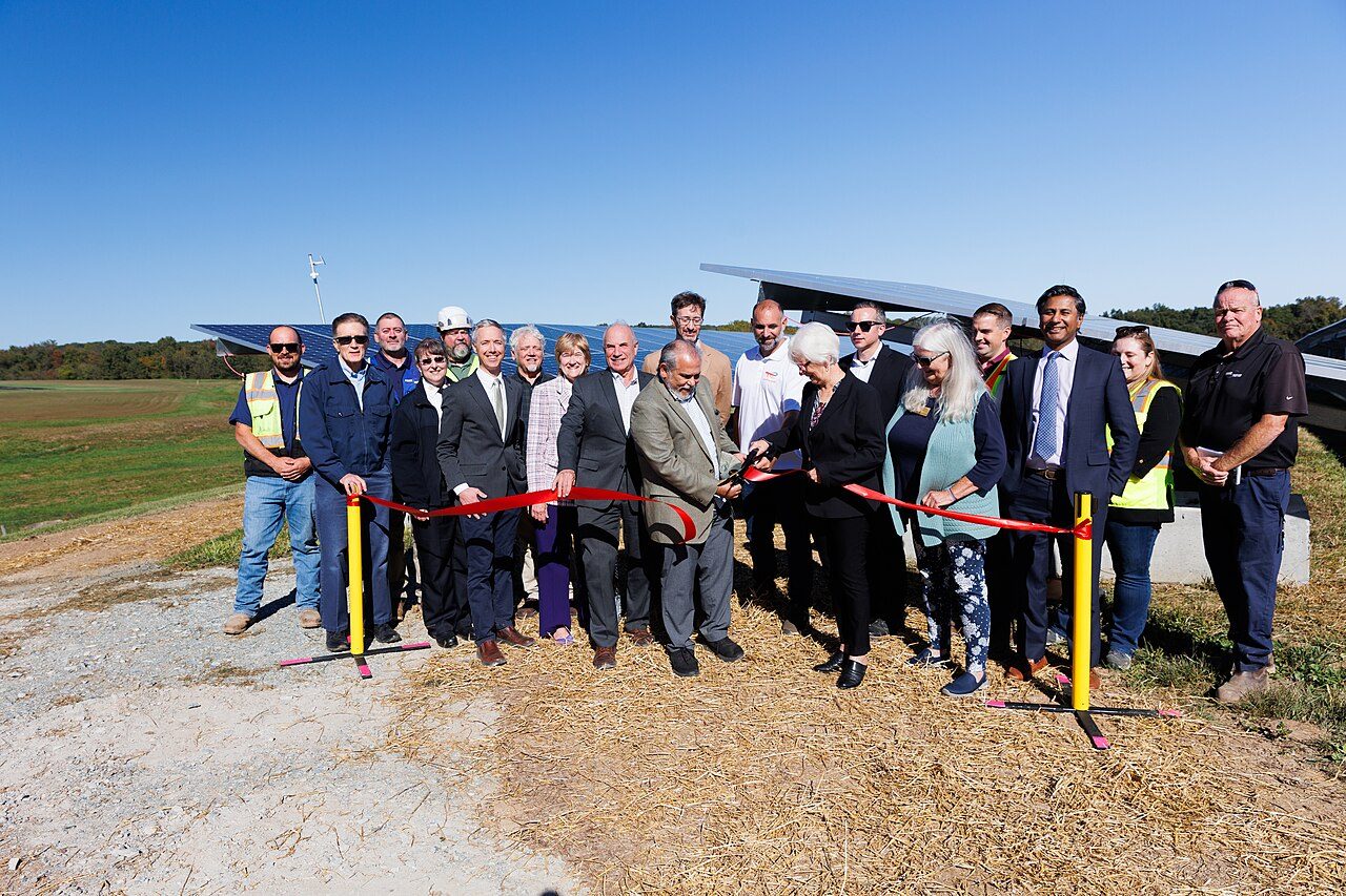 About twenty people, some in suits and some in high-visibility vests, cut a red ribbon in front of tilted ground-mounted solar panels on a grassy site under a clear blue sky.