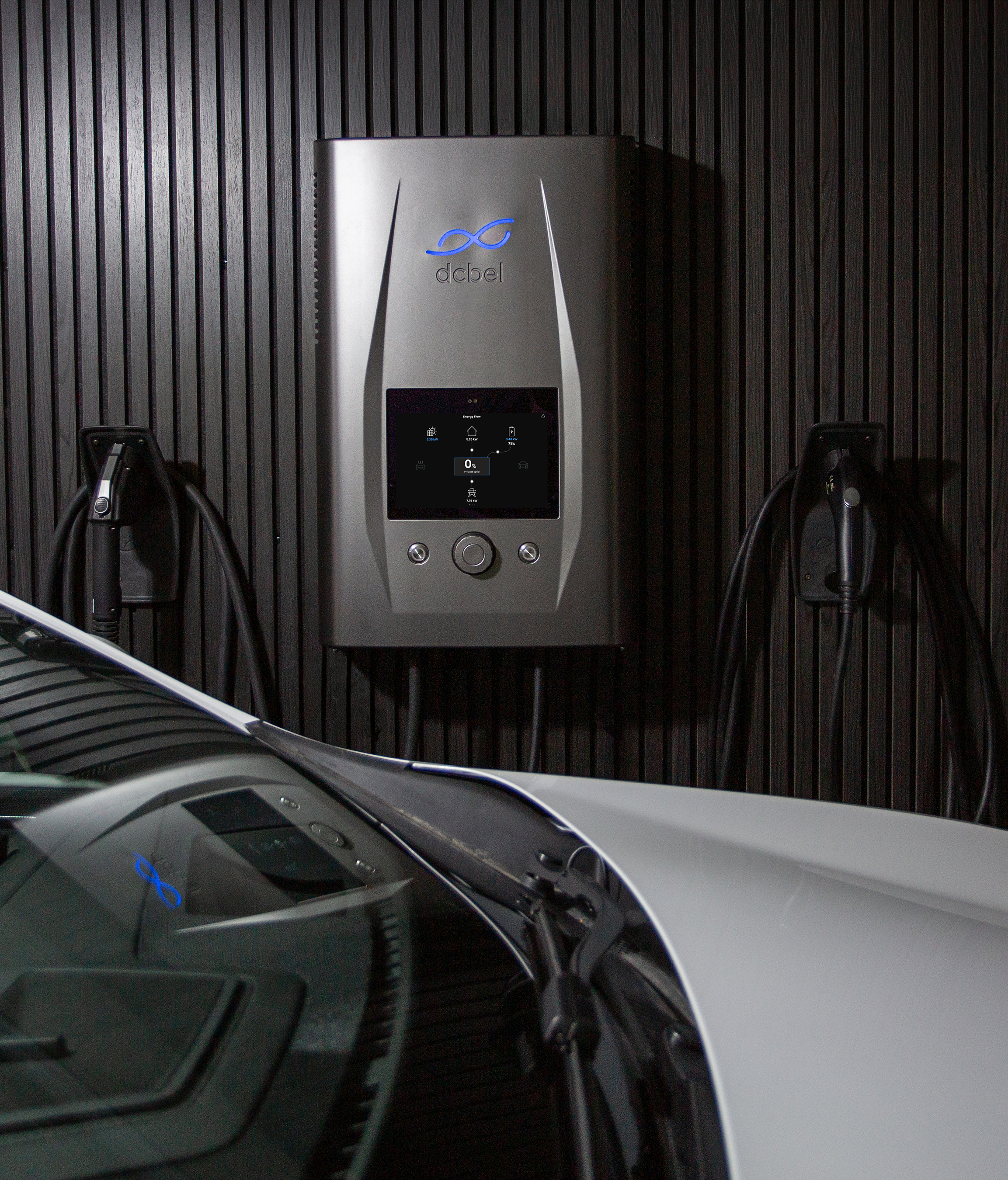 A wall-mounted dcbel Ara home energy station with its energy-flow screen lit, two holstered charging connectors, and the hood of a white electric car in the foreground