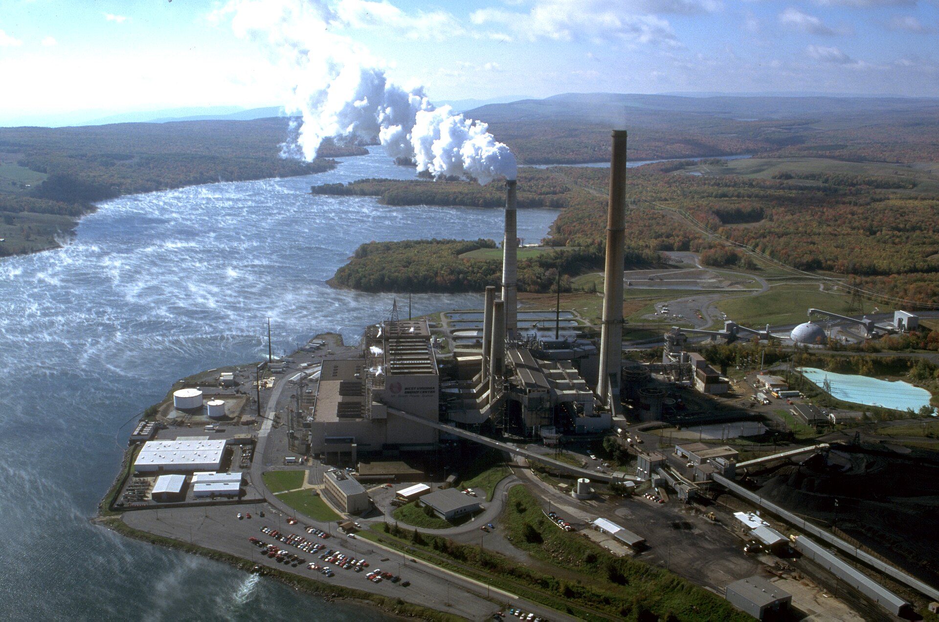 Aerial view of a coal power plant with tall smokestacks and rising steam on the shore of a lake, surrounded by autumn forest