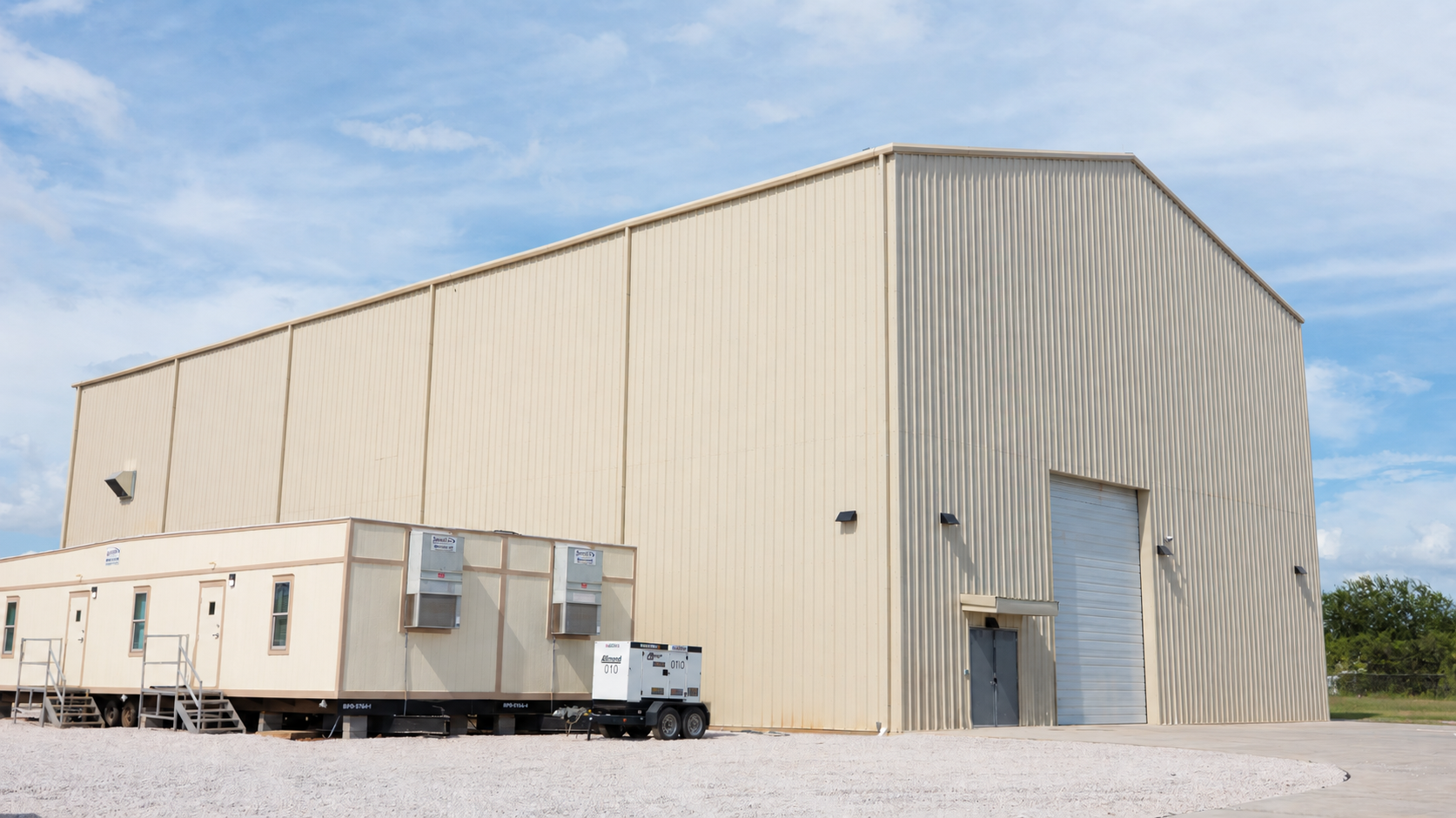 A tall beige metal-clad building with a large roll-up door and a side entrance, beside a single-story modular office trailer and a towable generator on a gravel lot under a partly cloudy sky.