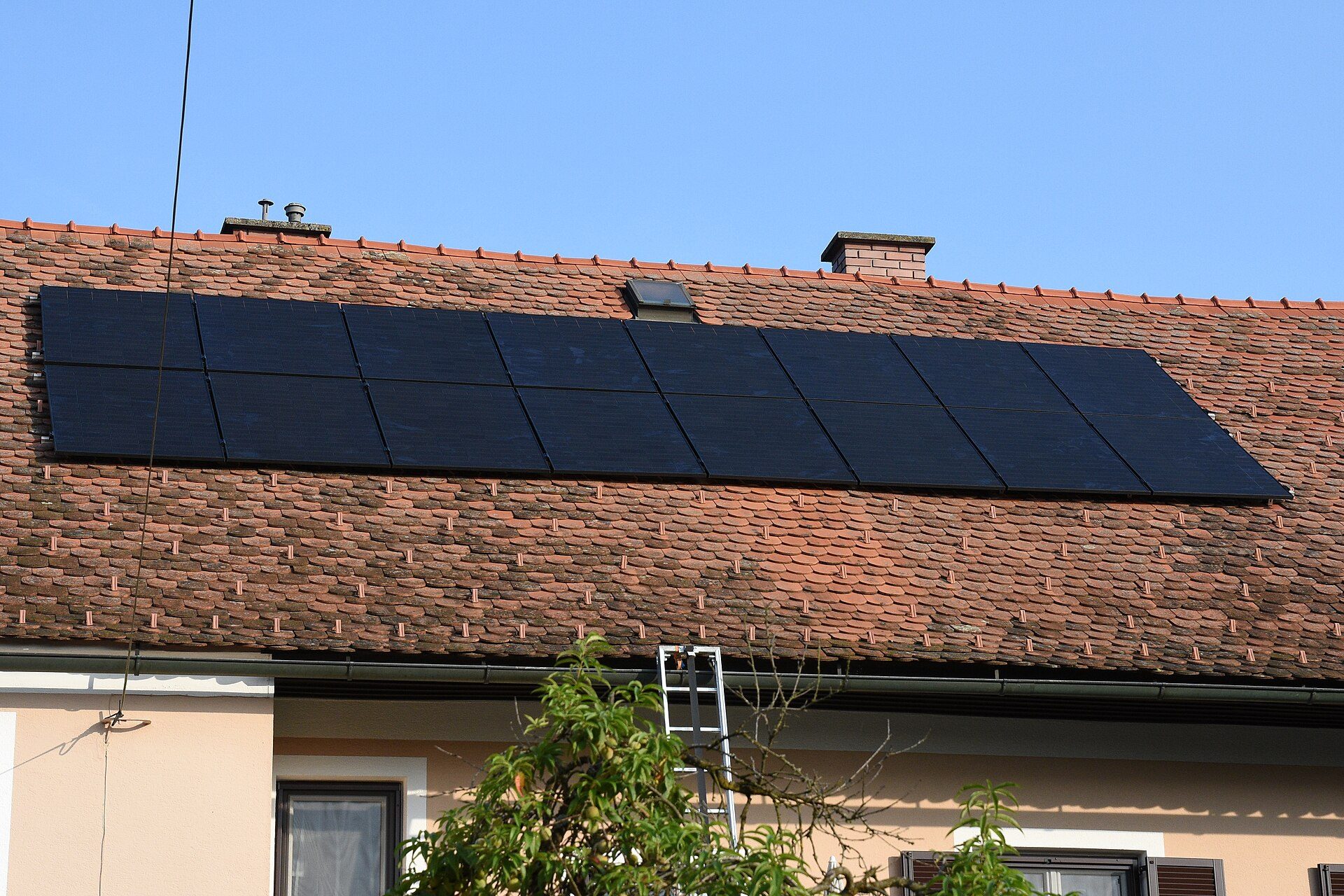 Two rows of black solar panels on a red clay-tile house roof, with a ladder leaning on the gutter and a fruit tree in the foreground
