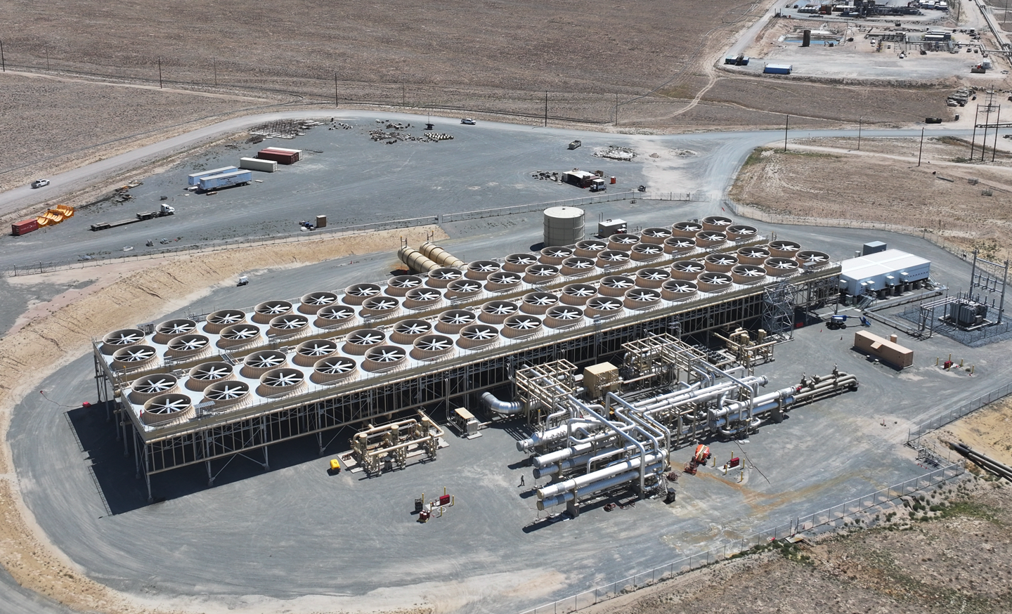 Aerial view of Fervo Energy's first GeoBlock at Cape Station in Beaver County, Utah: a long rectangular air-cooled condenser bank topped with rows of large fans, with steel piping, a storage tank and a small switchyard beside it on flat, bare ground