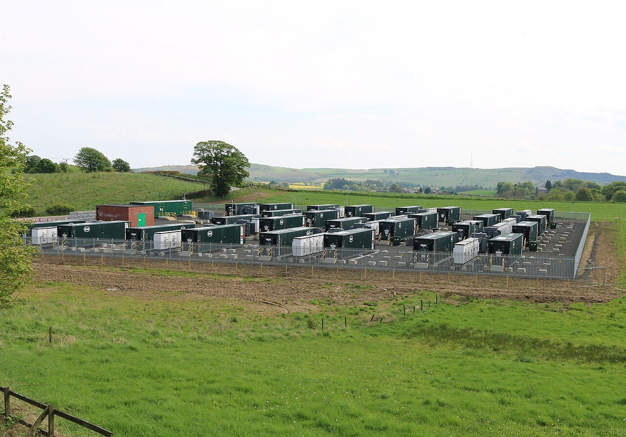 Rows of dark green battery enclosures and white equipment cabinets inside a fenced gravel compound set in green fields, with a large tree and low hills behind