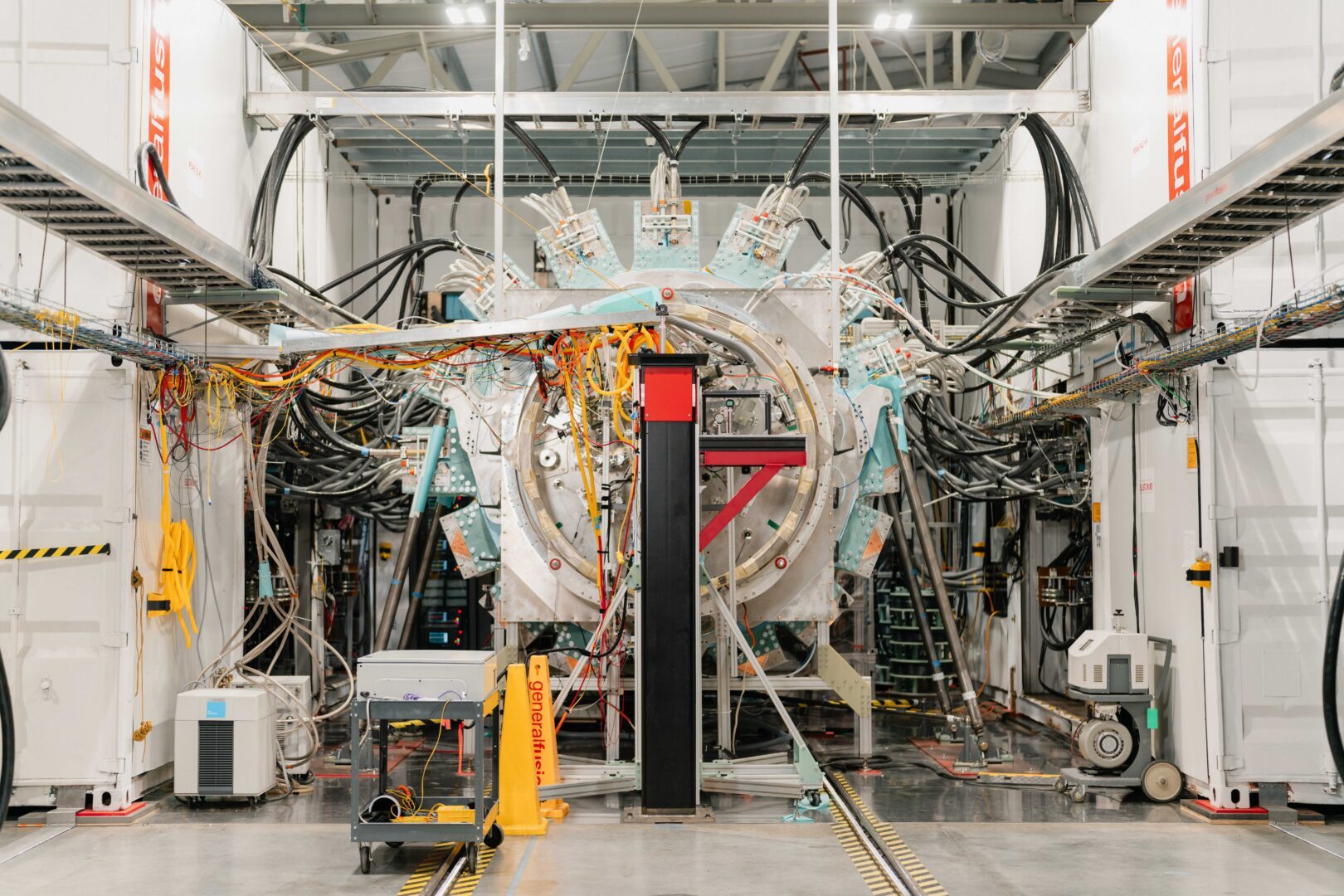 General Fusion's LM26 machine: a large circular steel assembly ringed by teal brackets and bundles of black cable, with a black and red post, a wheeled cart and yellow safety cones on the floor in front