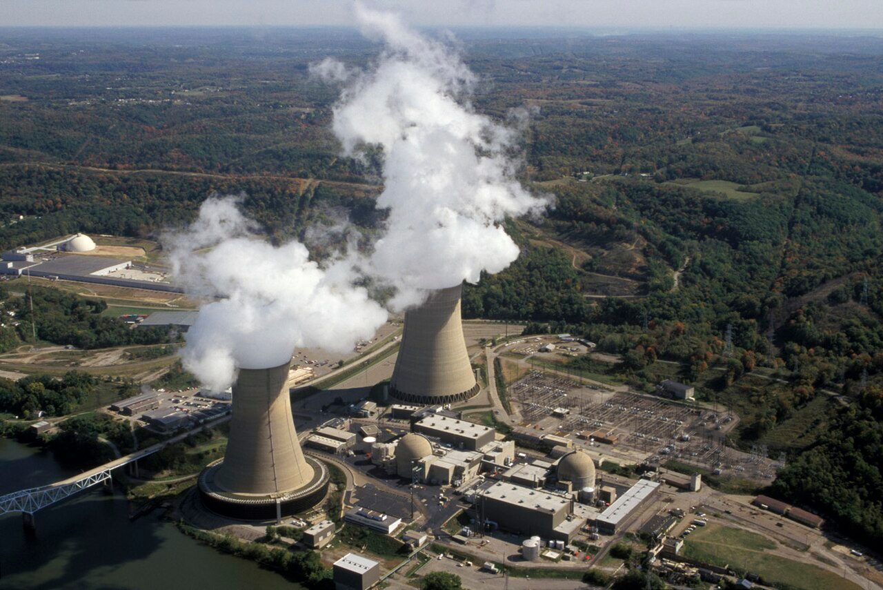 Aerial view of Beaver Valley Power Station: two cooling towers releasing steam plumes beside two domed reactor containment buildings, with a switchyard to the right, water and a bridge at lower left, and wooded hills behind.
