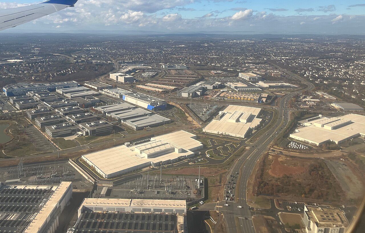 Aerial view from an airliner window of large white-roofed data center buildings and electrical substations beside a divided road near Ashburn, Virginia, with housing developments stretching to the horizon