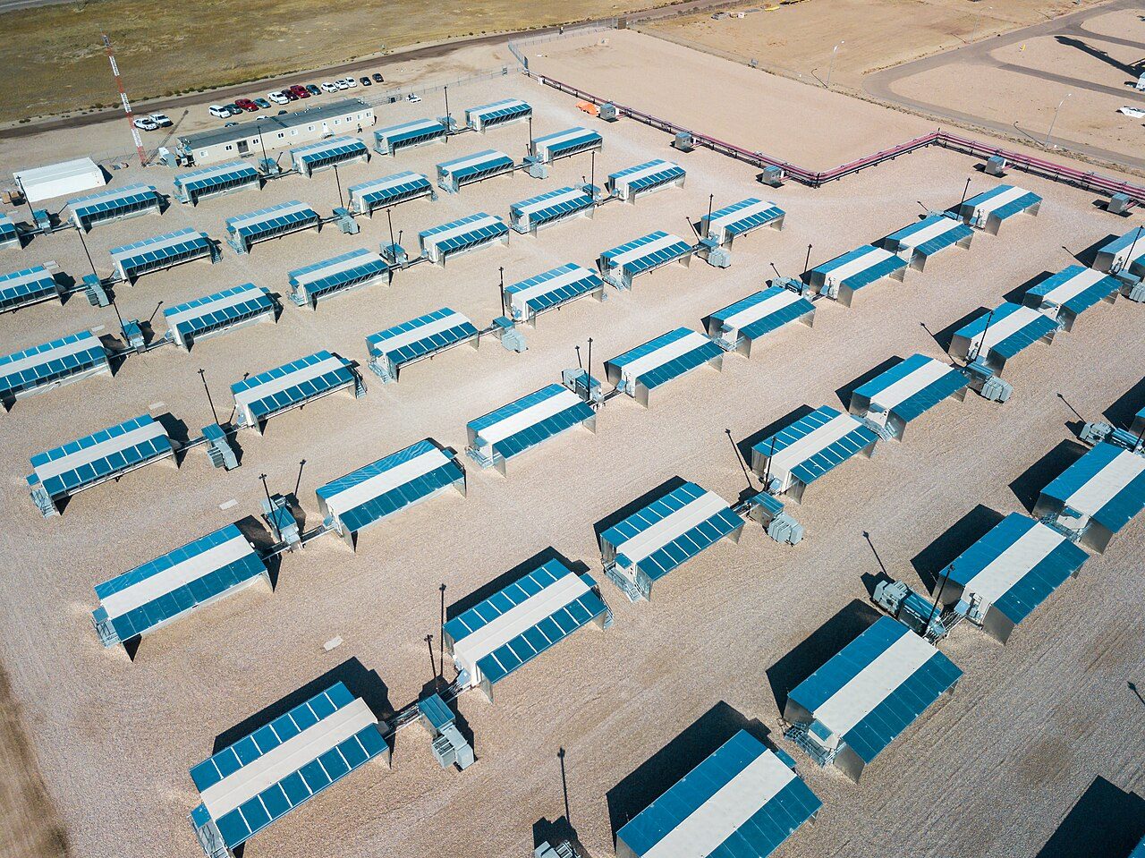 Aerial view of rows of blue-and-white mining containers on a gravel pad, each with a small transformer beside it, with a site office and parked cars at the far edge