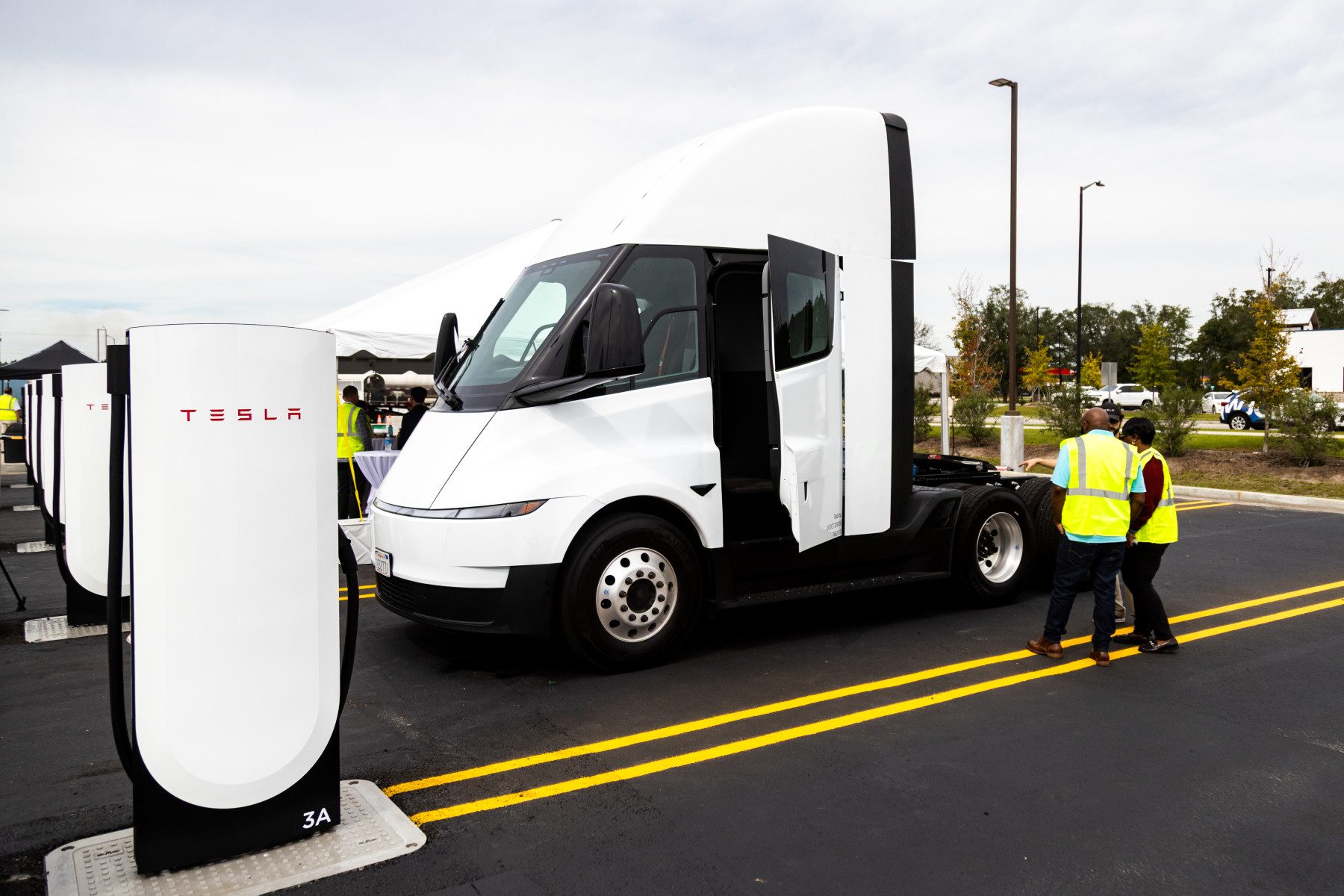 A white Tesla Semi tractor with a cab door open parked beside a row of white Tesla charging posts, the nearest marked 3A, as two people in high-visibility vests stand beside the truck
