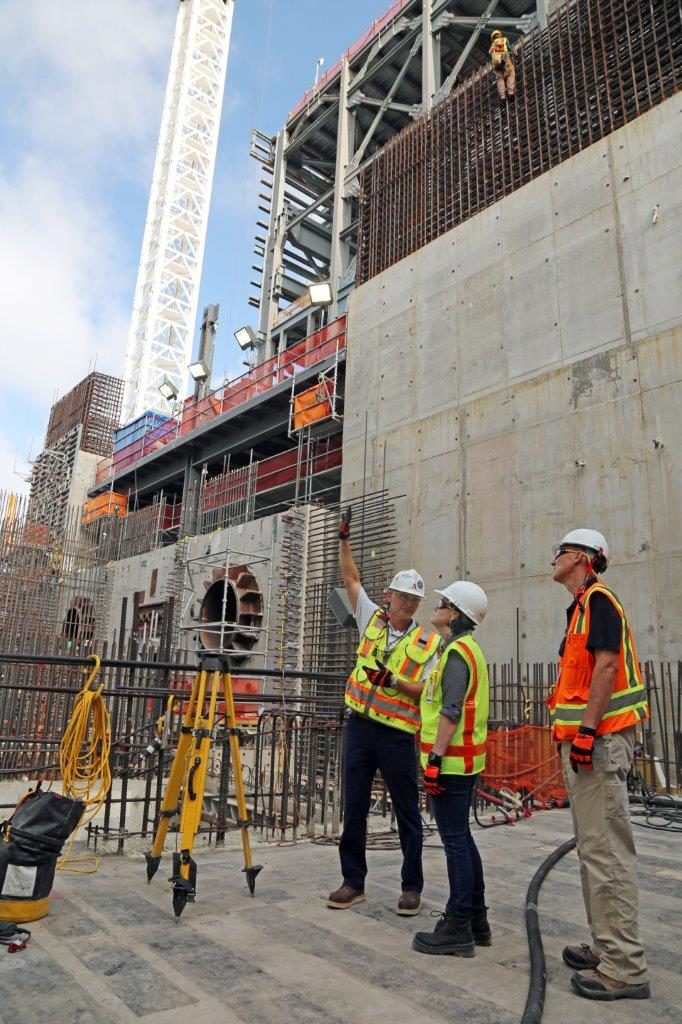 Three people in hard hats and high-visibility vests stand on a concrete deck at a reactor construction site; one points up at a tall concrete wall studded with rebar, beneath a steel frame and a white lattice crane tower against a blue sky.