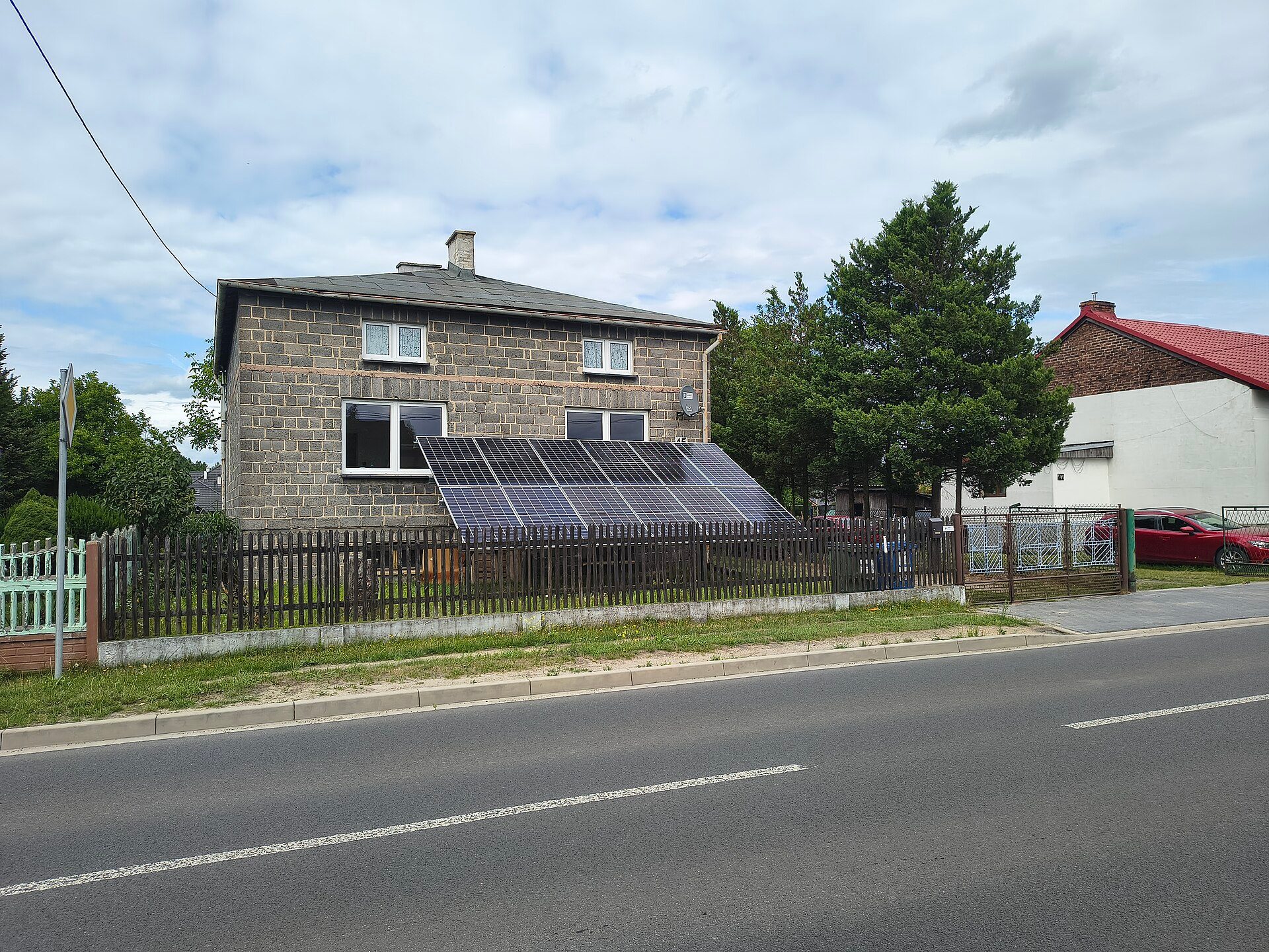 A two-storey block house in Bojszowy, Poland, with two rows of solar panels on a tilted frame in its front yard behind a wooden picket fence, beside a road